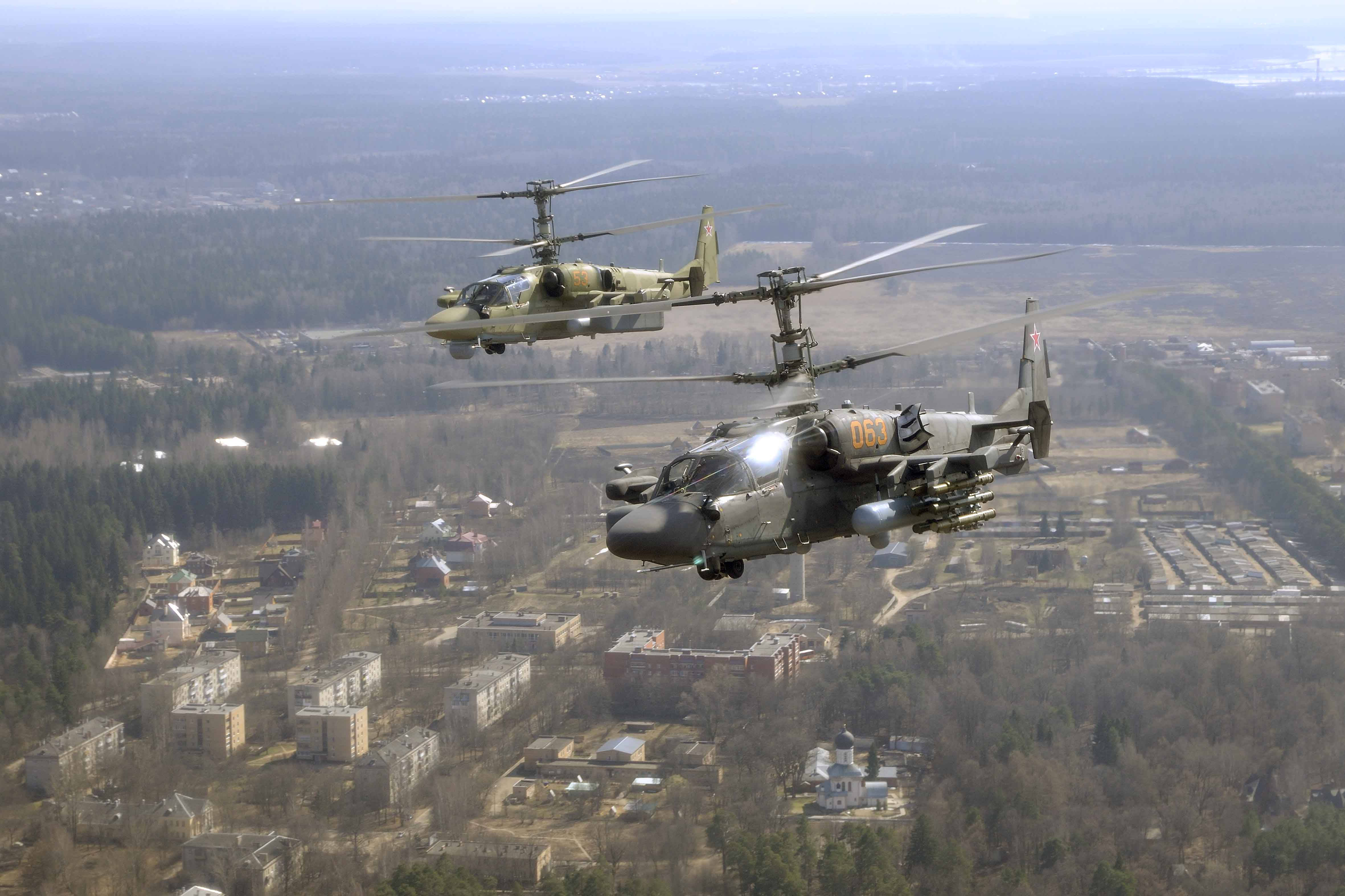 Helicopter 桌布 壁紙 — 兩架軍用直升機以編隊飛行，飛越一個坐落於光禿樹木和遠處森林之間的廣闊城鎮。