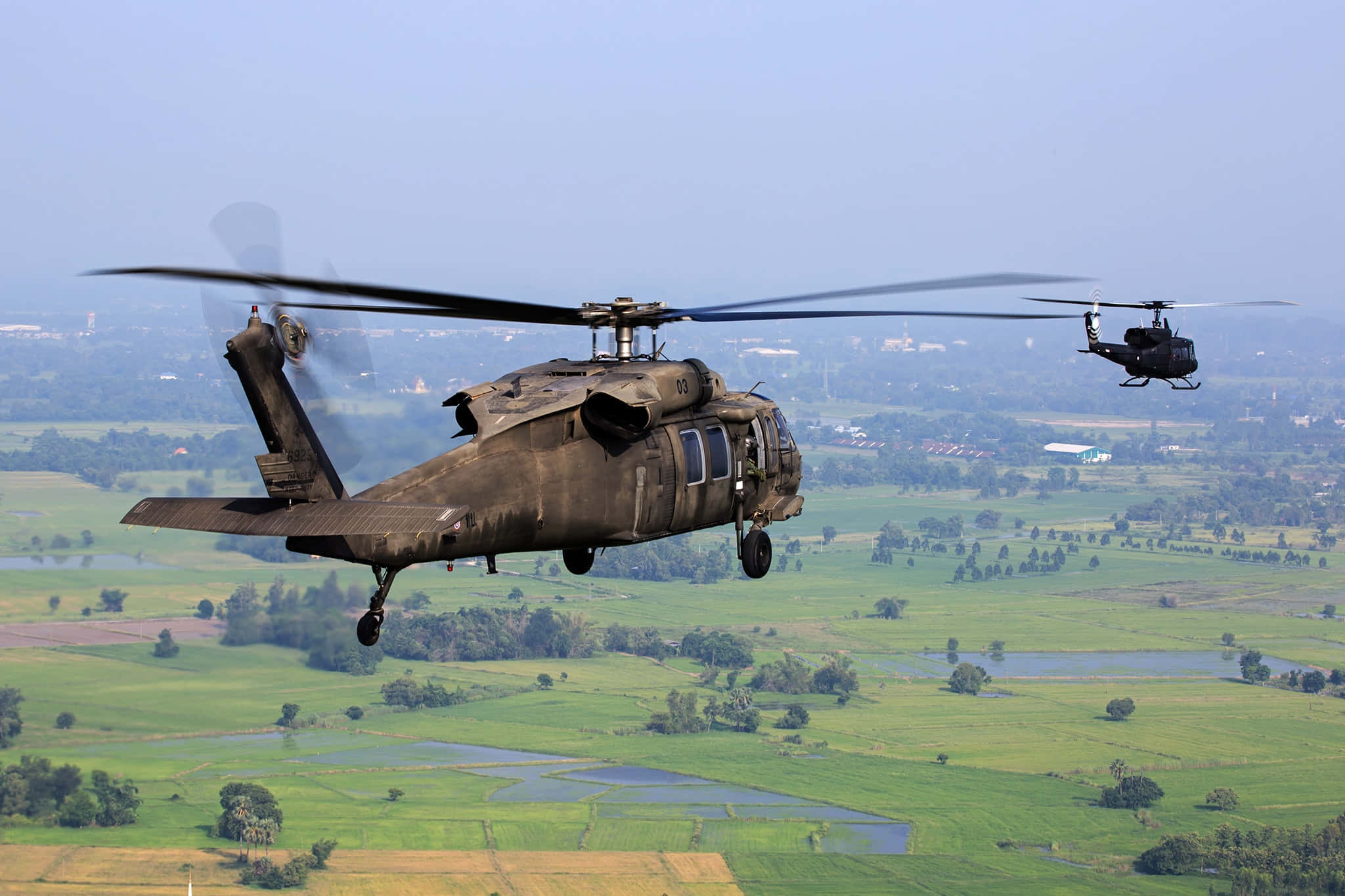 Helicopter 桌布 壁紙 — 兩架軍用直升機在朦朧的天空下低飛過一片綠油油的鄉村，有稻田和零散的樹木。
