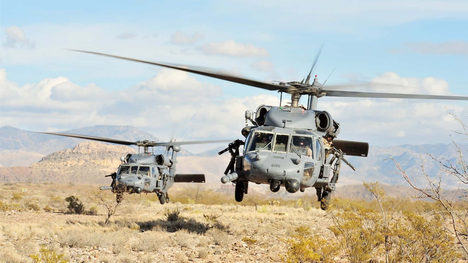 Helicopter 桌布 壁紙 — 兩架軍用直升機在部分多雲的天空下，低空盤旋於乾燥的沙漠景觀之上，遠方有山脈。
