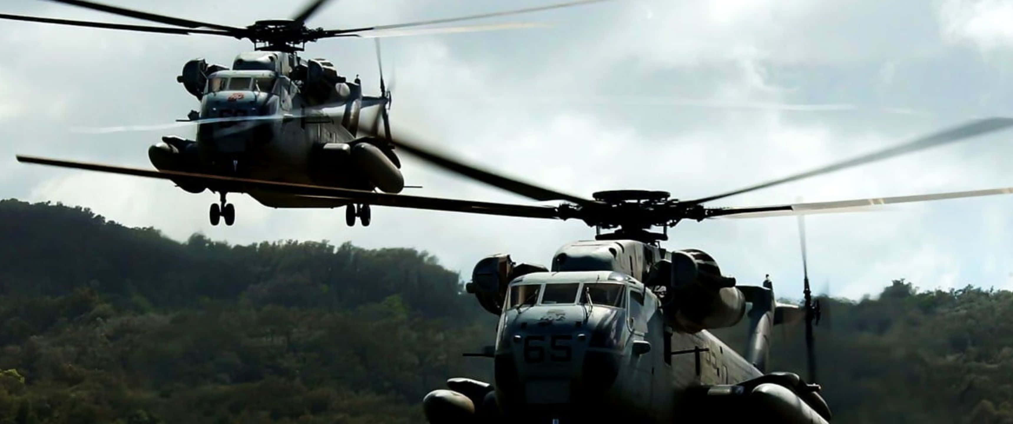 3440x1440p Helicopters 桌布 壁紙 — 兩架軍用直升機在陰天籠罩下盤旋於樹木繁茂的山坡上空。