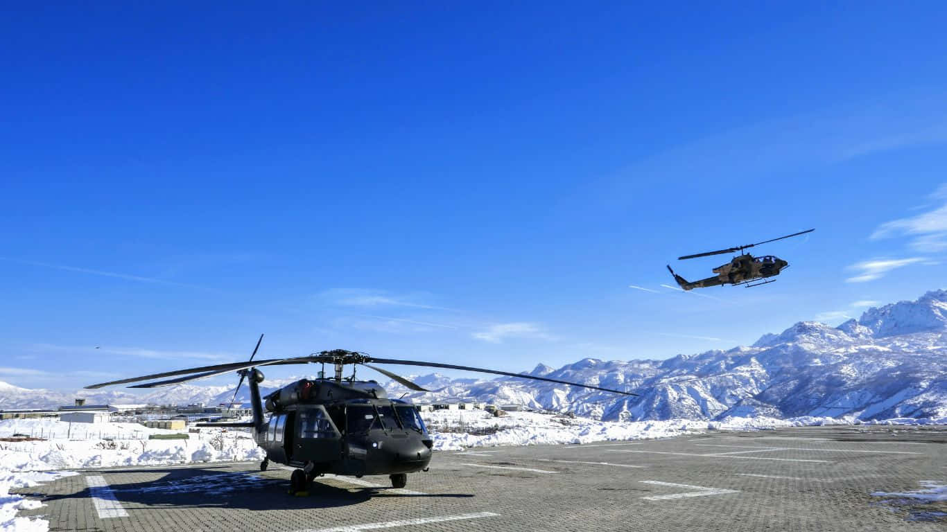 1366x768 Helicopters 桌布 壁紙 — 兩架黑色軍用直升機，一架停放在積雪的停機坪上，另一架在晴朗的藍天中飛行，背景是白雪覆蓋的山脈。