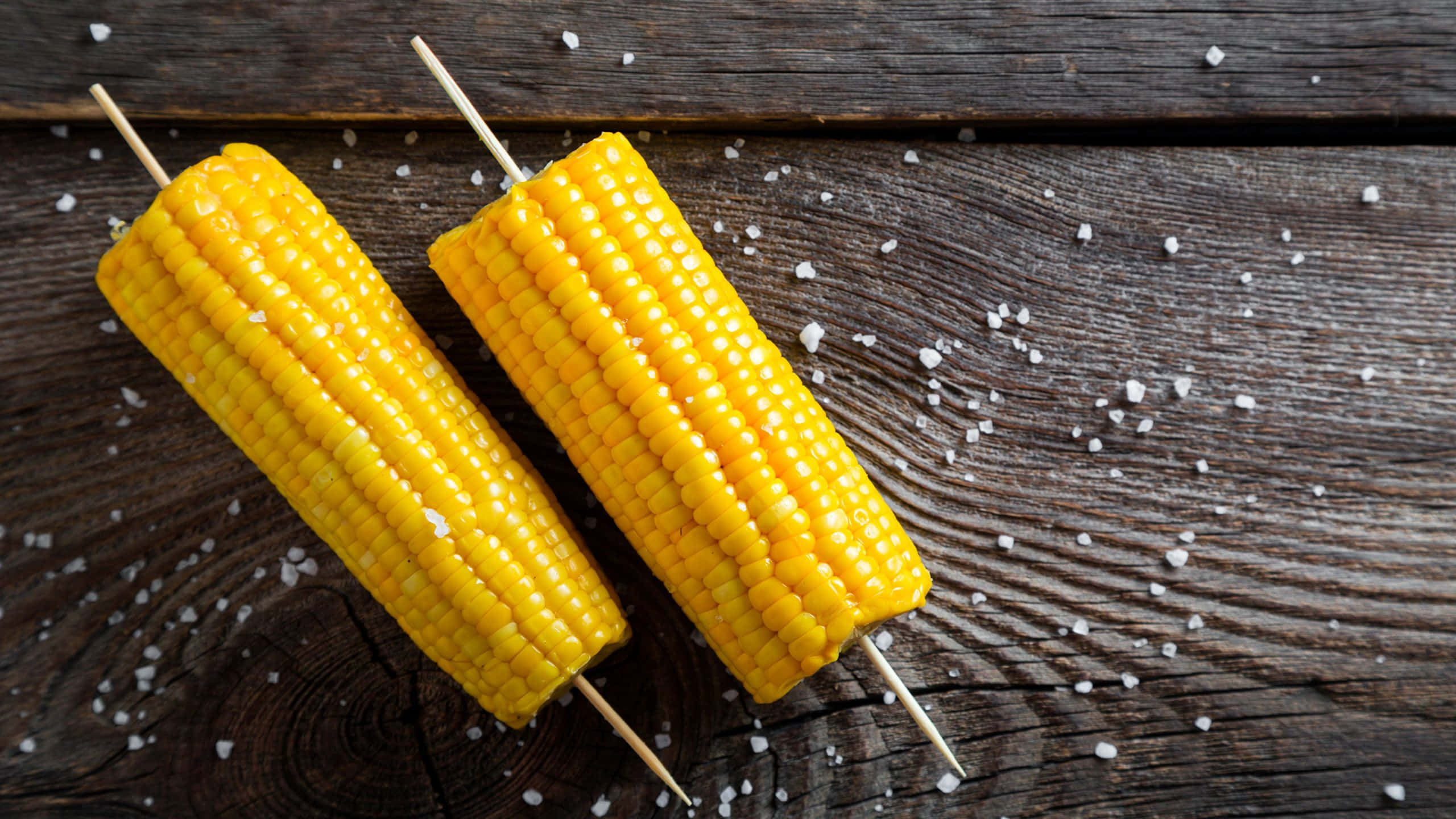 Corn 桌布 壁紙 — 兩根鹽水玉米串放在經風化的木質表面上，周圍散落著粗鹽。