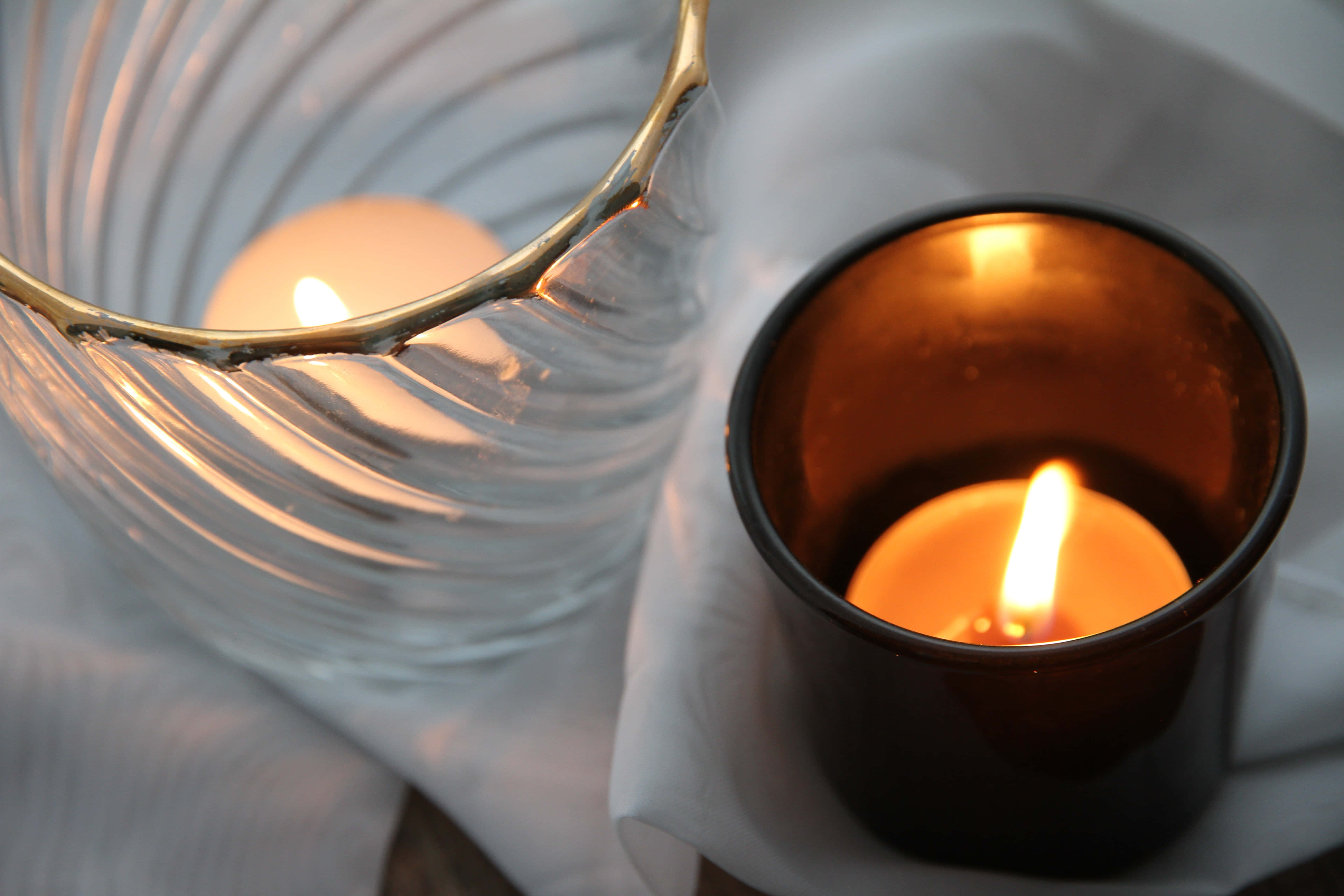 Calming 桌布 壁紙 — 兩根點燃的蠟燭溫暖地發光，一根在帶棱紋的玻璃花瓶裡，另一根在深琥珀色的罐子裡，放在柔軟的白色布料上。