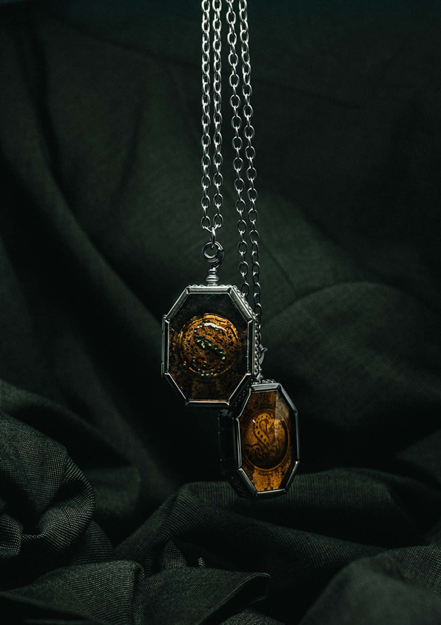 Horcrux 桌布 壁紙 — 兩條八角形吊墜項鍊，帶有精緻的金色龍紋設計，懸掛在深色紋理布料背景上。