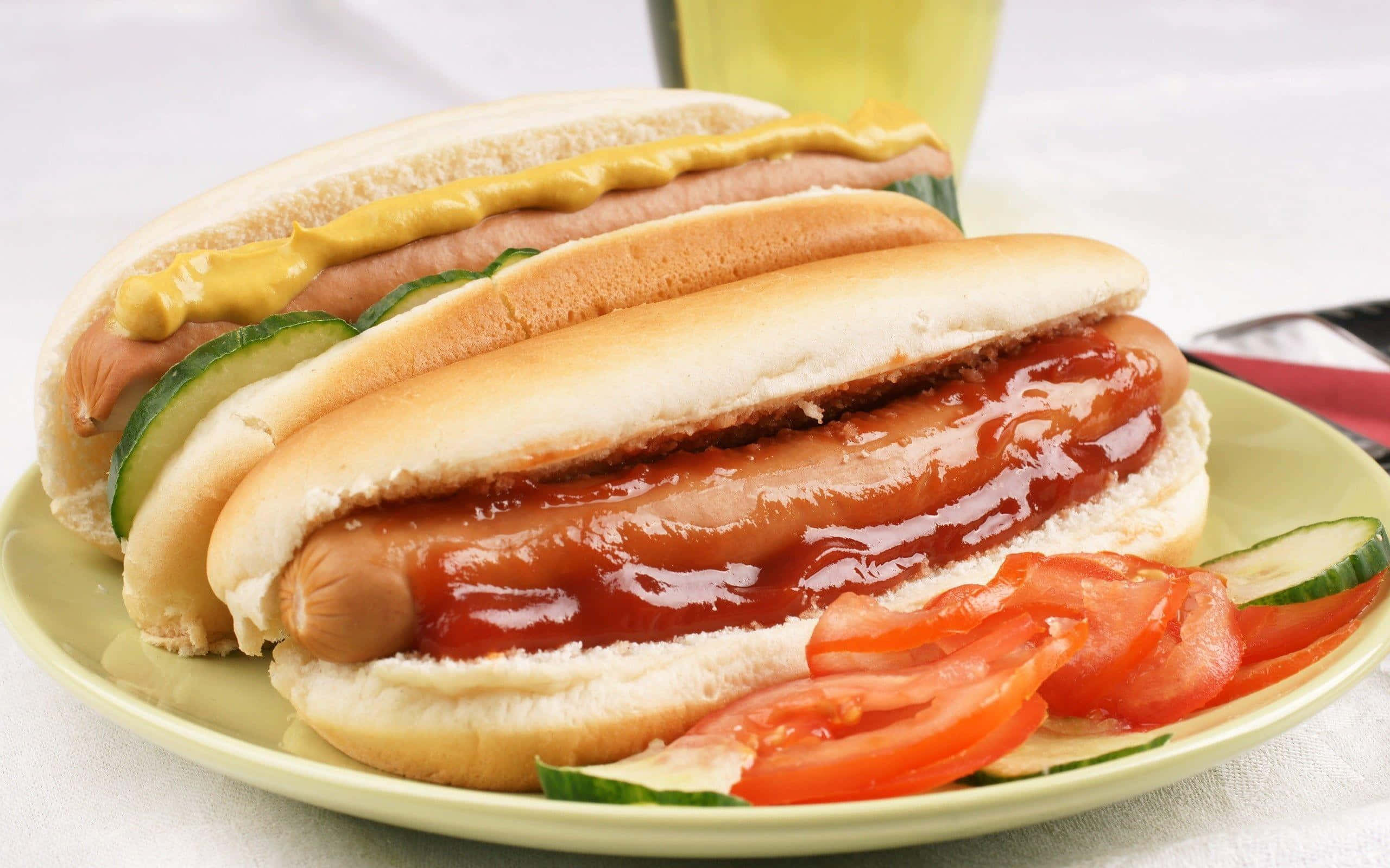 Hot Dog 桌布 壁紙 — 兩條多汁的熱狗，上面淋上番茄醬、芥末和新鮮蔬菜，放在綠色盤子上，背景有一杯飲料。