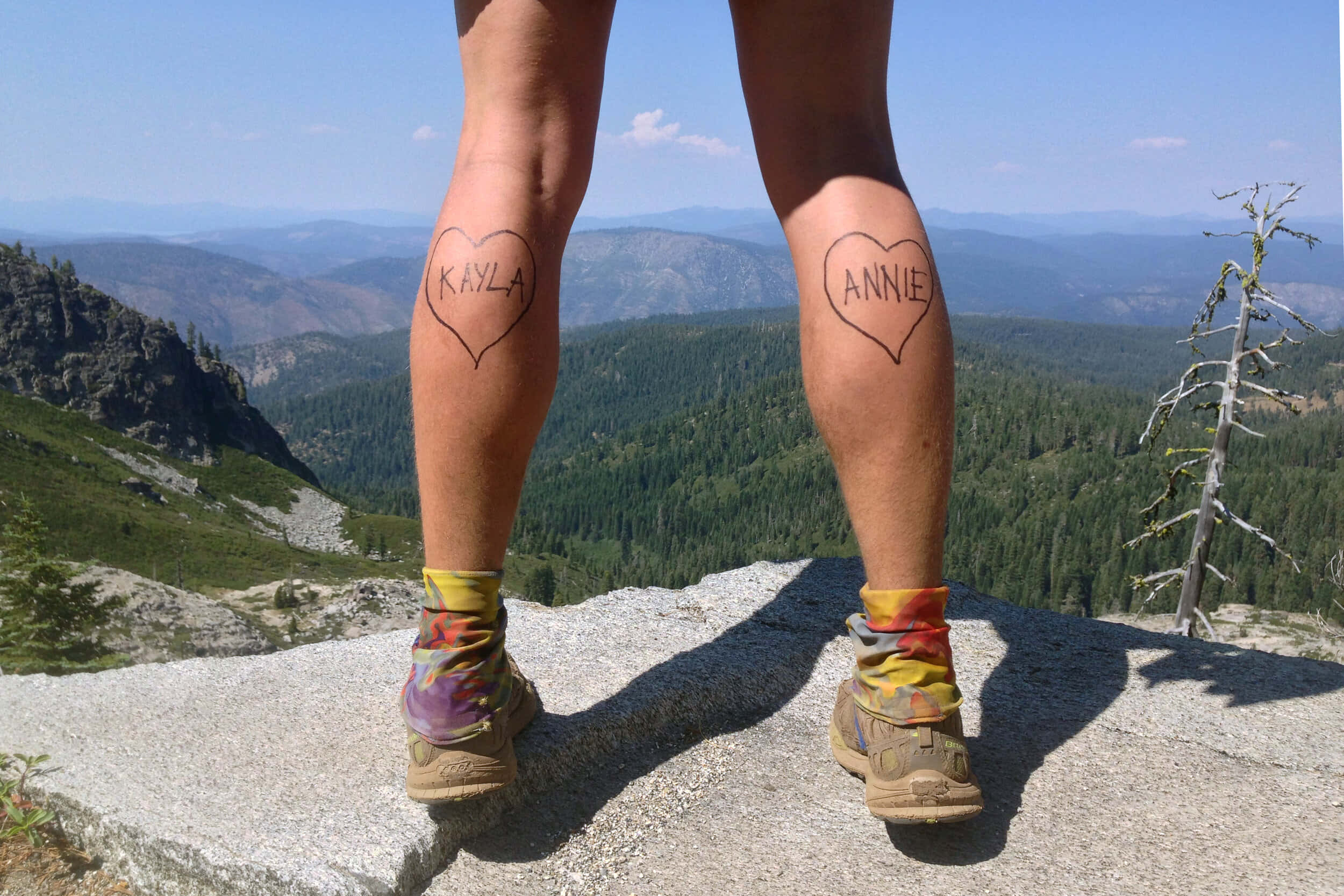 Hiking 桌布 壁紙 — 兩條帶有心形刺青、寫著 Kayla 和 Annie 的腿，站在岩石山頂上，俯瞰著晴朗藍天下廣闊的森林山谷。
