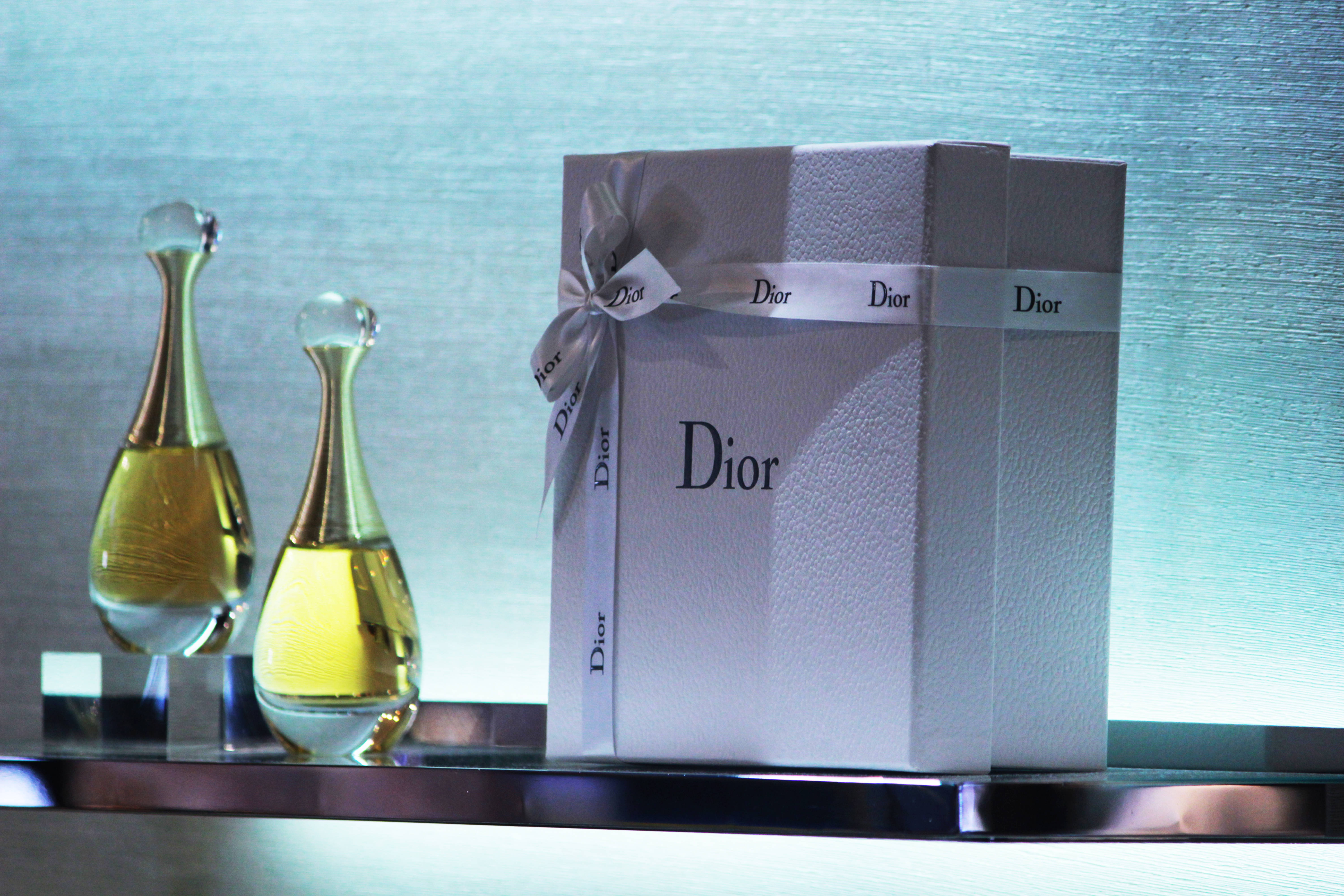Dior 動態 桌布 壁紙 — 兩款優雅的Dior香水瓶在帶有紋理的藍色背景下，在一個用絲帶繫著的銀色禮物盒旁邊閃閃發光。
