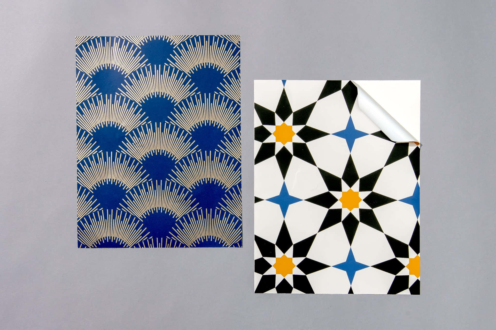 Tiles 桌布 壁紙 — 兩款幾何壁紙樣品，帶有醒目的藍色、金色和黑白圖案，平放在灰色表面上，其中一張部分剝離。