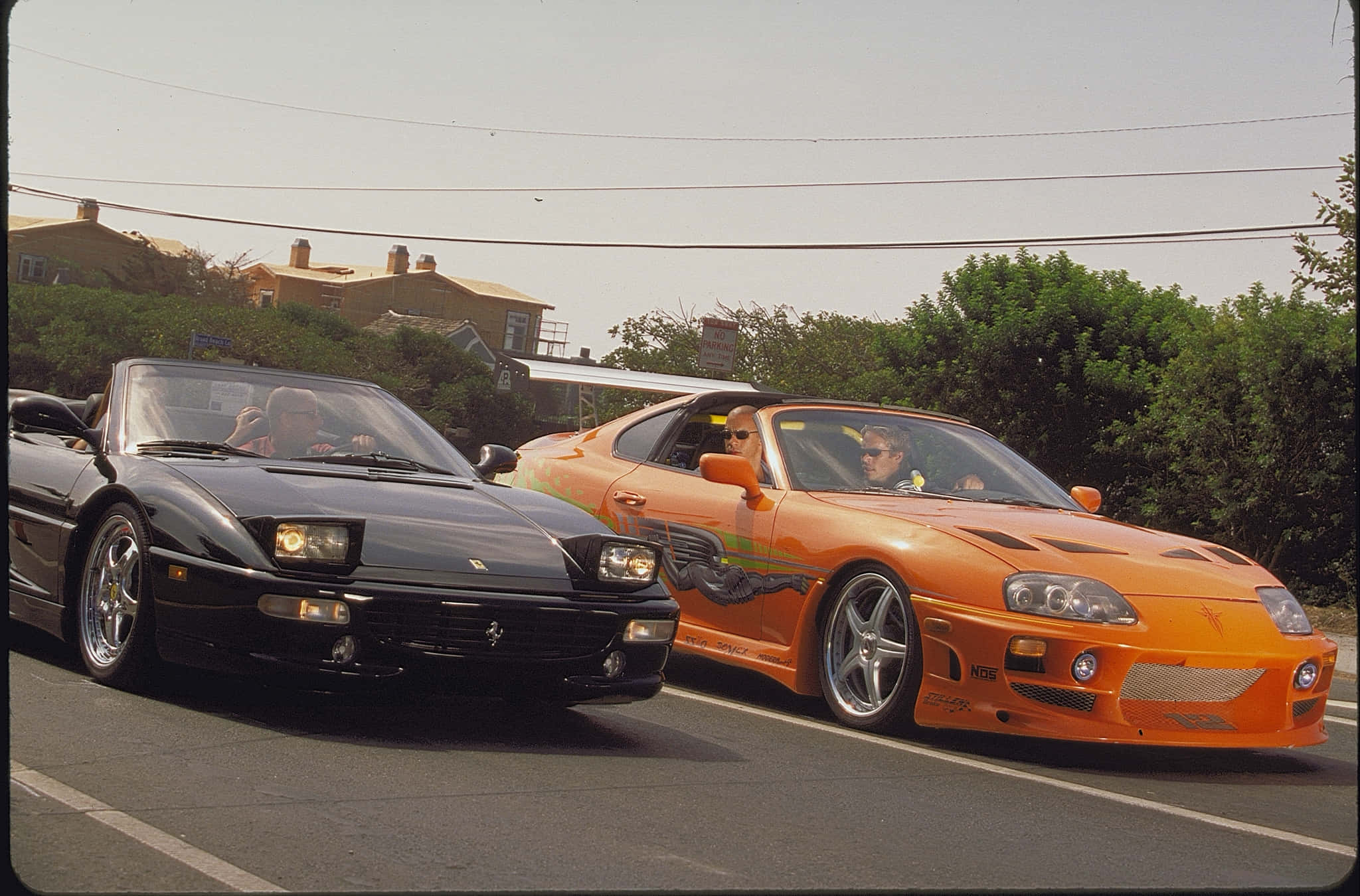 Fast And Furious 1 桌布 壁紙 — 兩款標誌性跑車，黑色法拉利和橙色豐田Supra，在陽光明媚的郊區道路上並肩競速。