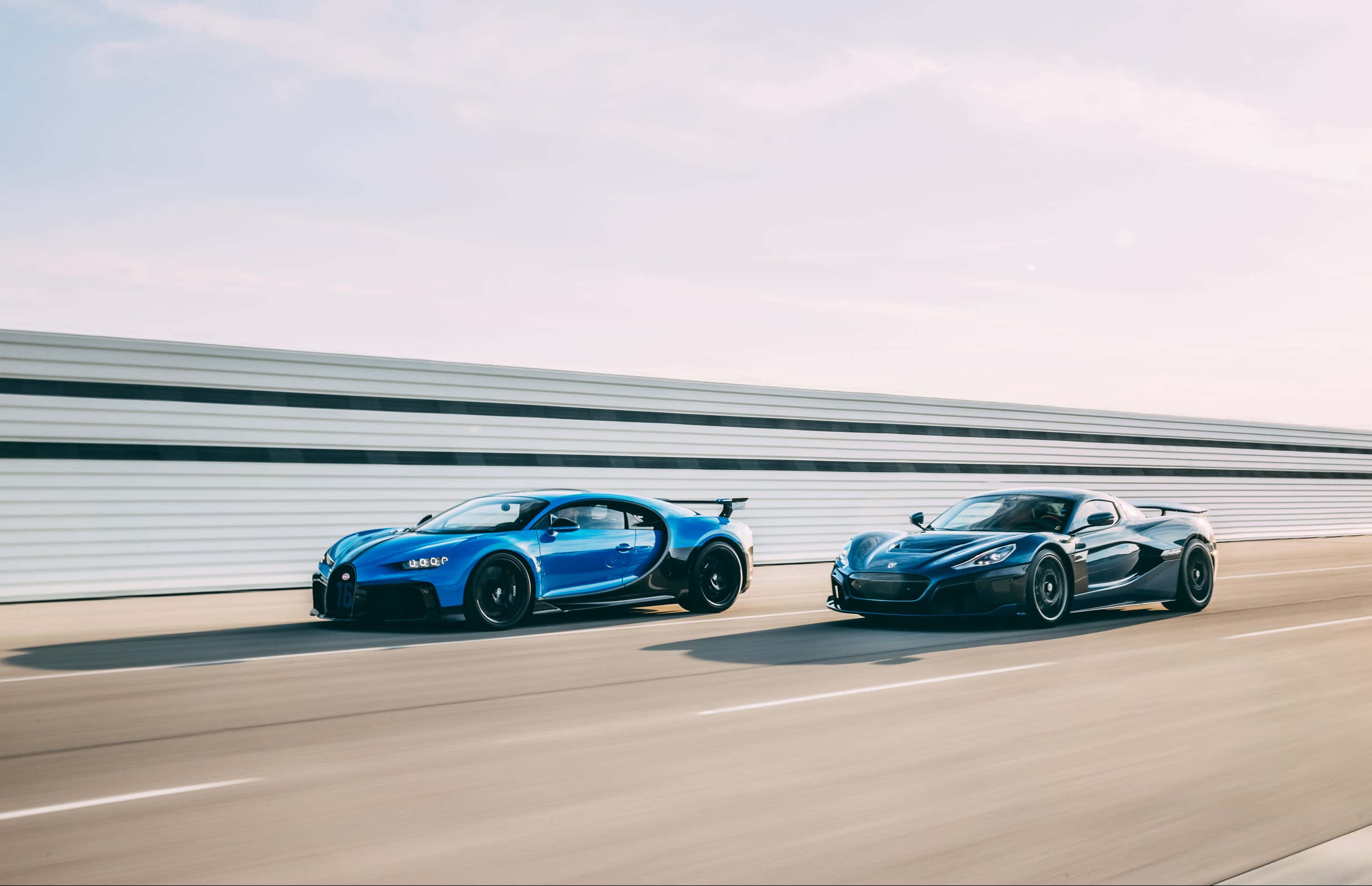 Rimac 桌布 壁紙 — 兩款高性能超級跑車，一輛充滿活力的藍色布加迪和一輛時尚的黑色麥拿侖，在陽光明媚的賽道上並肩競速。