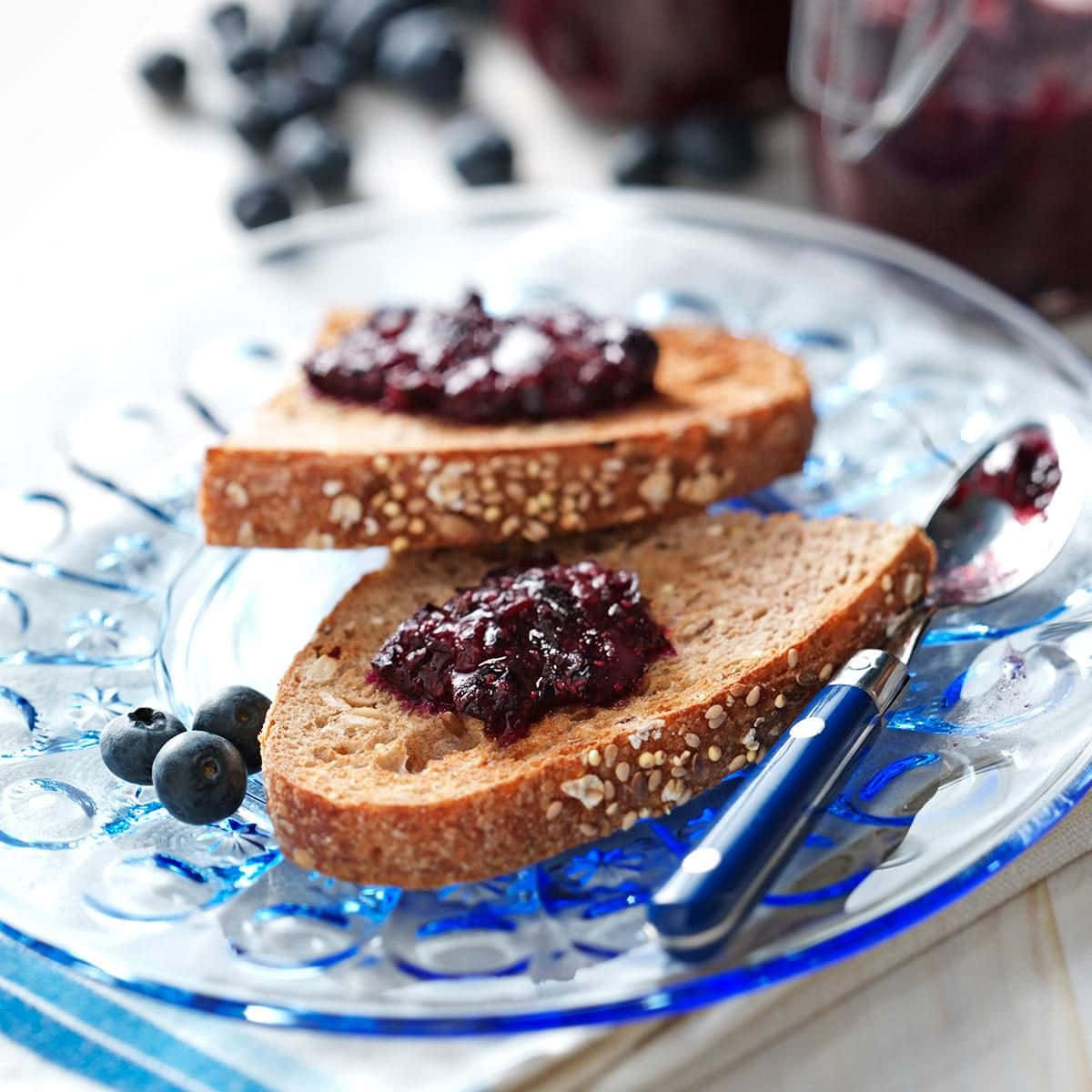 Blueberry Jam 桌布 壁紙 — 兩片籽粒全麥吐司，上面塗有光澤的藍莓果醬，放在華麗的藍色玻璃盤上，搭配新鮮漿果和一支湯匙。