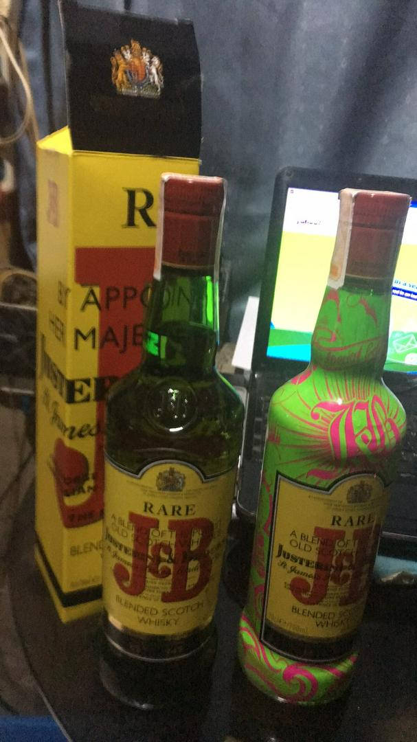drinks 桌布 壁紙 — 兩瓶 J&B Rare 威士忌，一瓶標準款，一瓶霓虹粉紅和綠色圖案款，放在深色表面上，旁邊是它們的盒子和一台筆記型電腦。