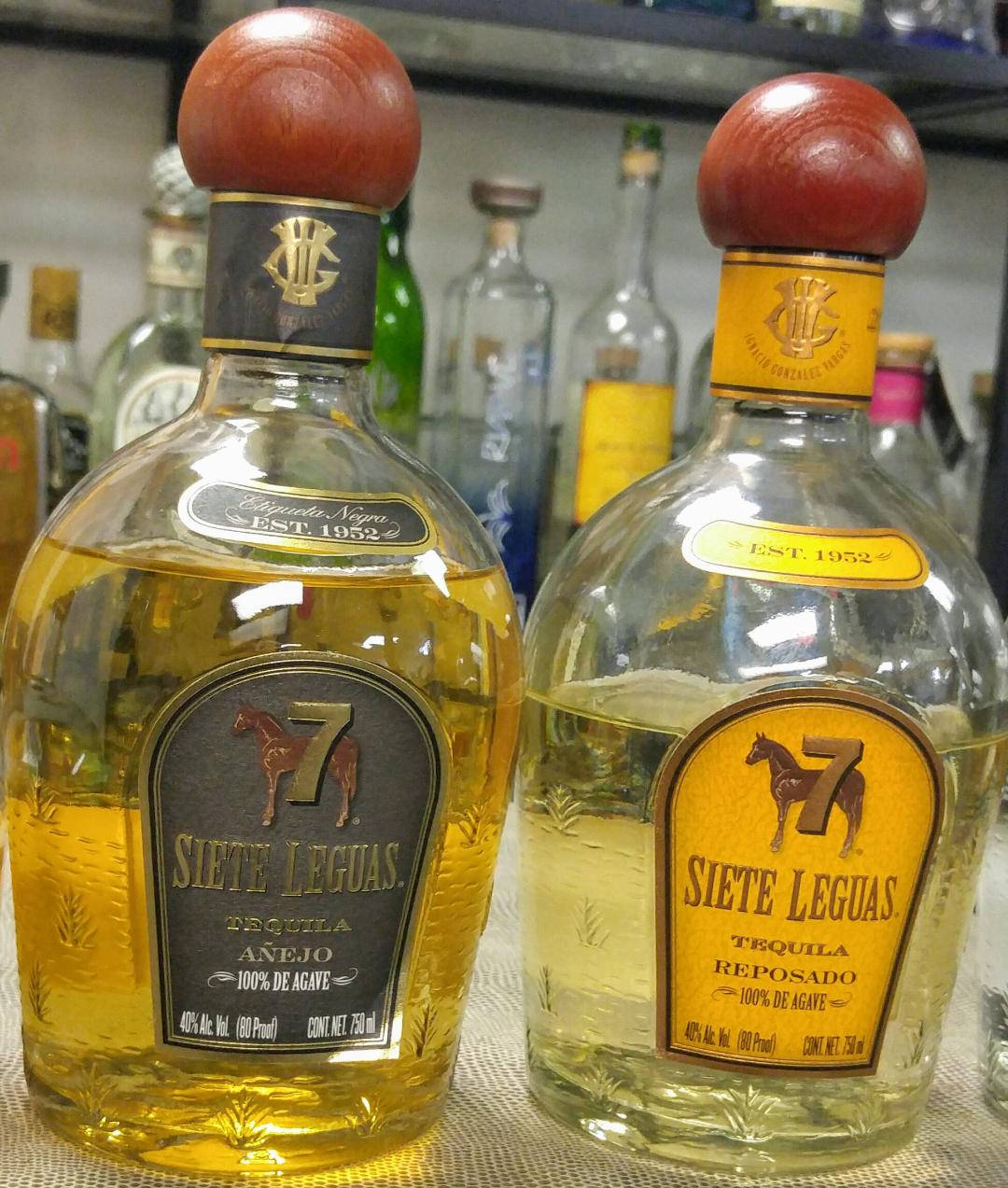 雙螢幕桌布 桌布 壁紙 — 兩瓶 Siete Leguas 龍舌蘭酒，一瓶 añejo 一瓶 reposado，並排放在架子上，背景有其他酒瓶。