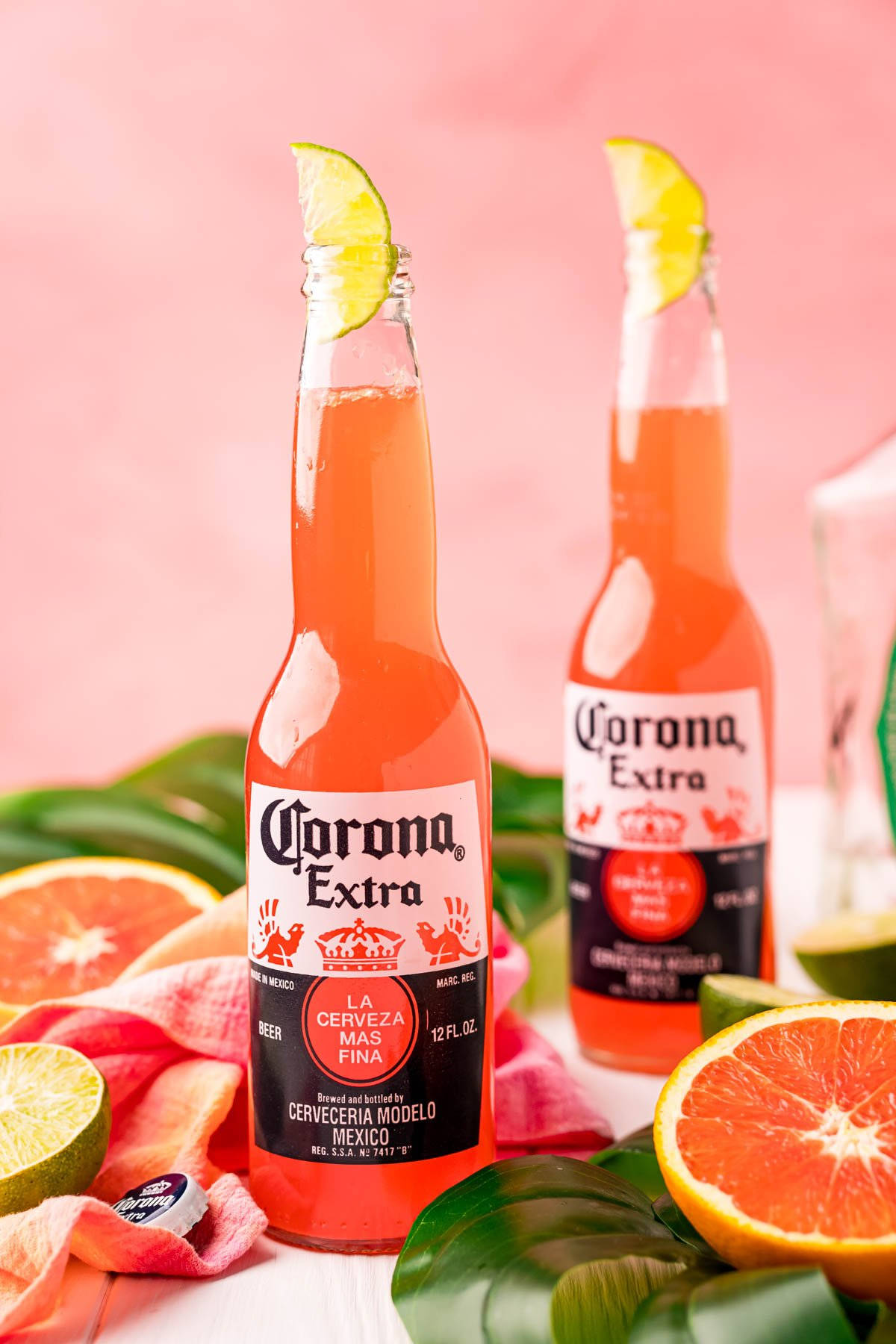 cocktail 桌布 壁紙 — 兩瓶Corona Extra啤酒配上萊姆角，放在切片柑橘和熱帶葉子之間，背景是柔和的粉紅色。