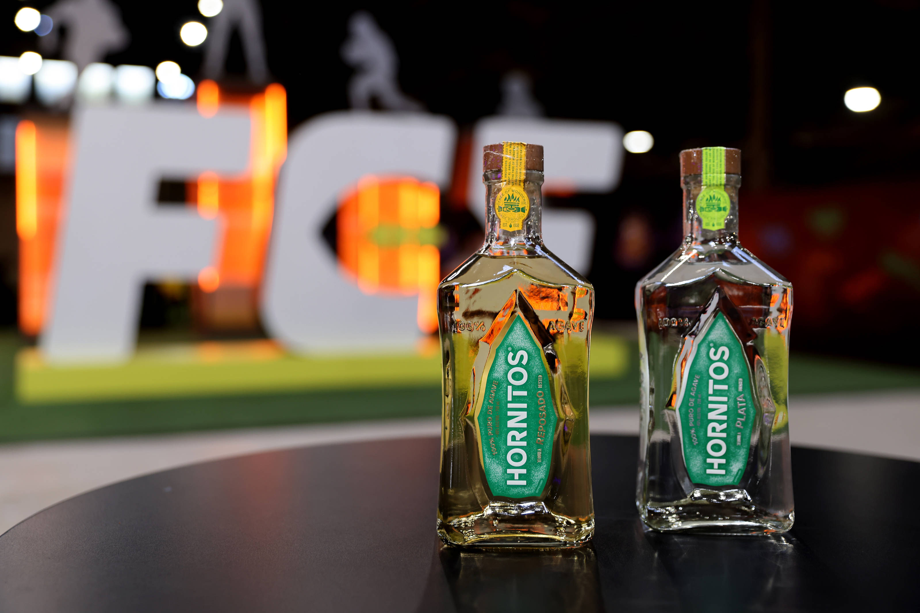 alcohol 桌布 壁紙 — 兩瓶Hornitos龍舌蘭酒在明亮燈光下於深色桌子上閃耀，模糊背景中有一個發光的「FG」標誌。