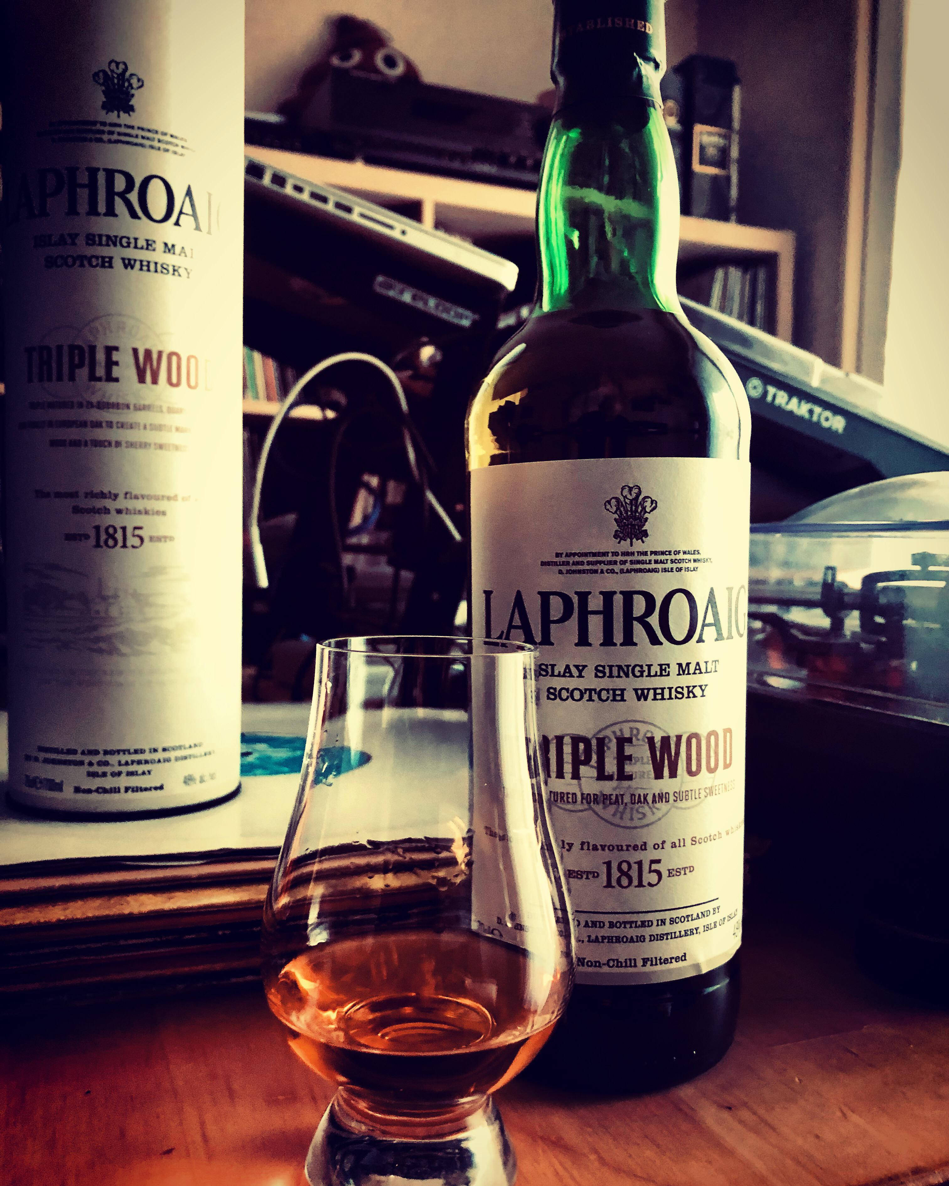 iPhone 桌布 壁紙 — 兩瓶Laphroaig Triple Wood威士忌和一個品酒杯放在木桌上，房間光線溫暖，背景有音響設備。