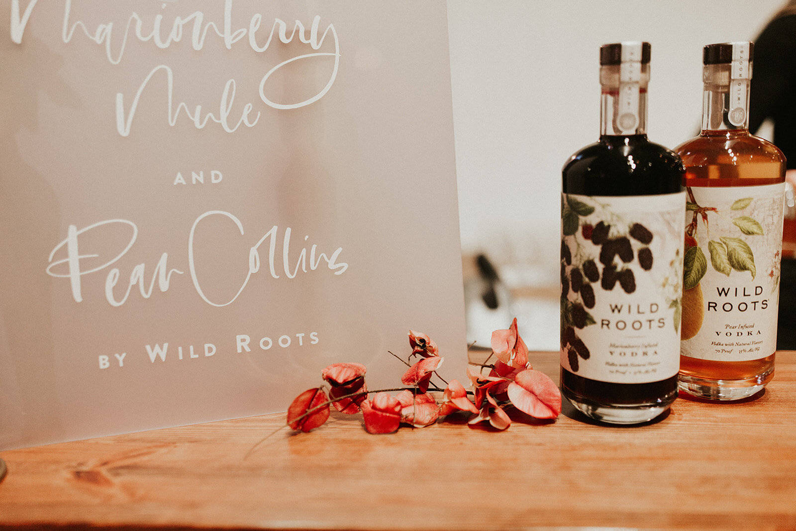 bottle 桌布 壁紙 — 兩瓶Wild Roots伏特加放在木製吧檯上，旁邊是Marionberry Mule和Pear Collins雞尾酒的標牌，並飾有乾燥的紅花。