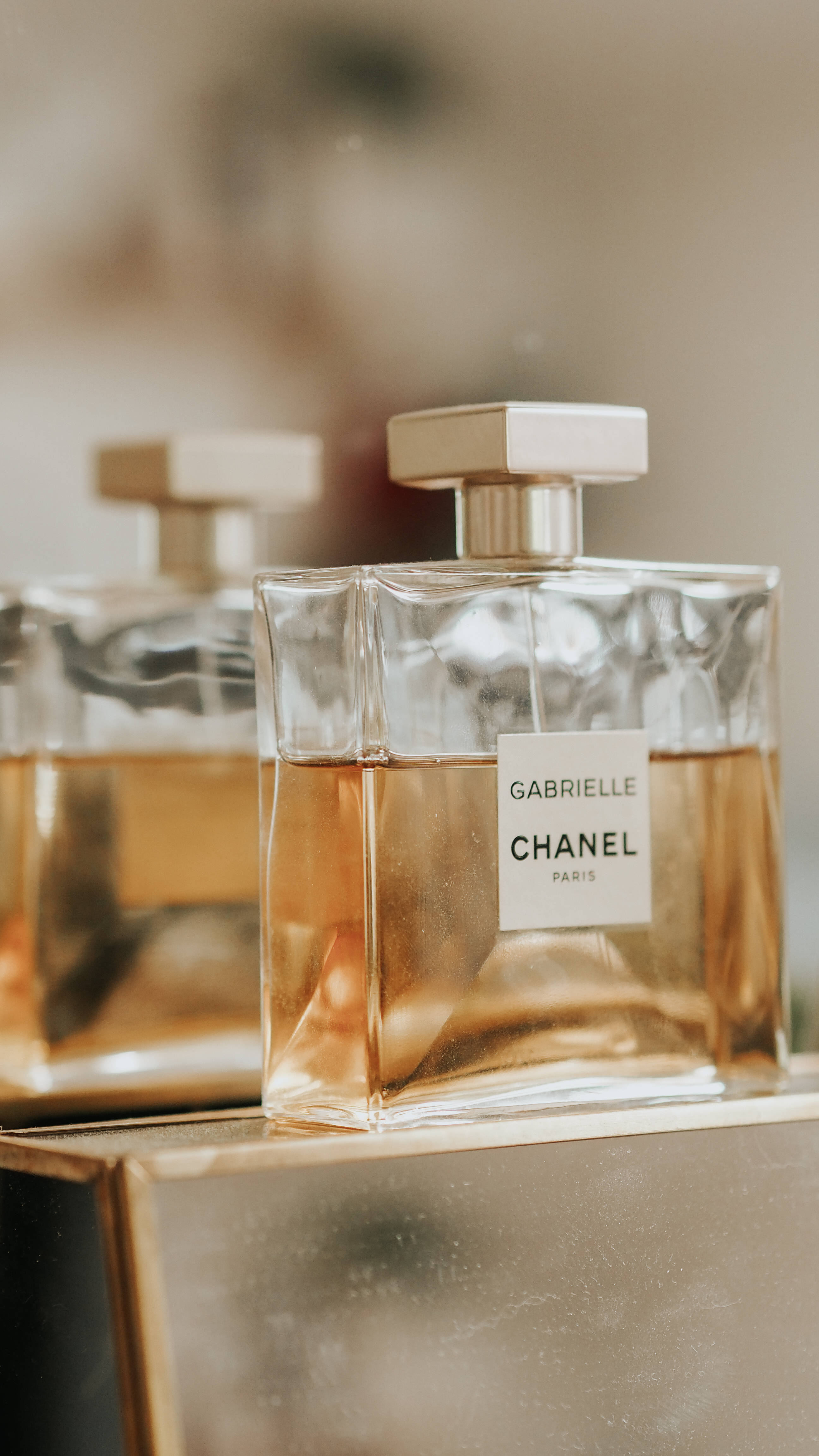 Chanel 桌布 壁紙 — 兩瓶優雅的 Chanel Gabrielle 香水，裝有琥珀色液體，放置在反射金色表面上，光線柔和溫暖。
