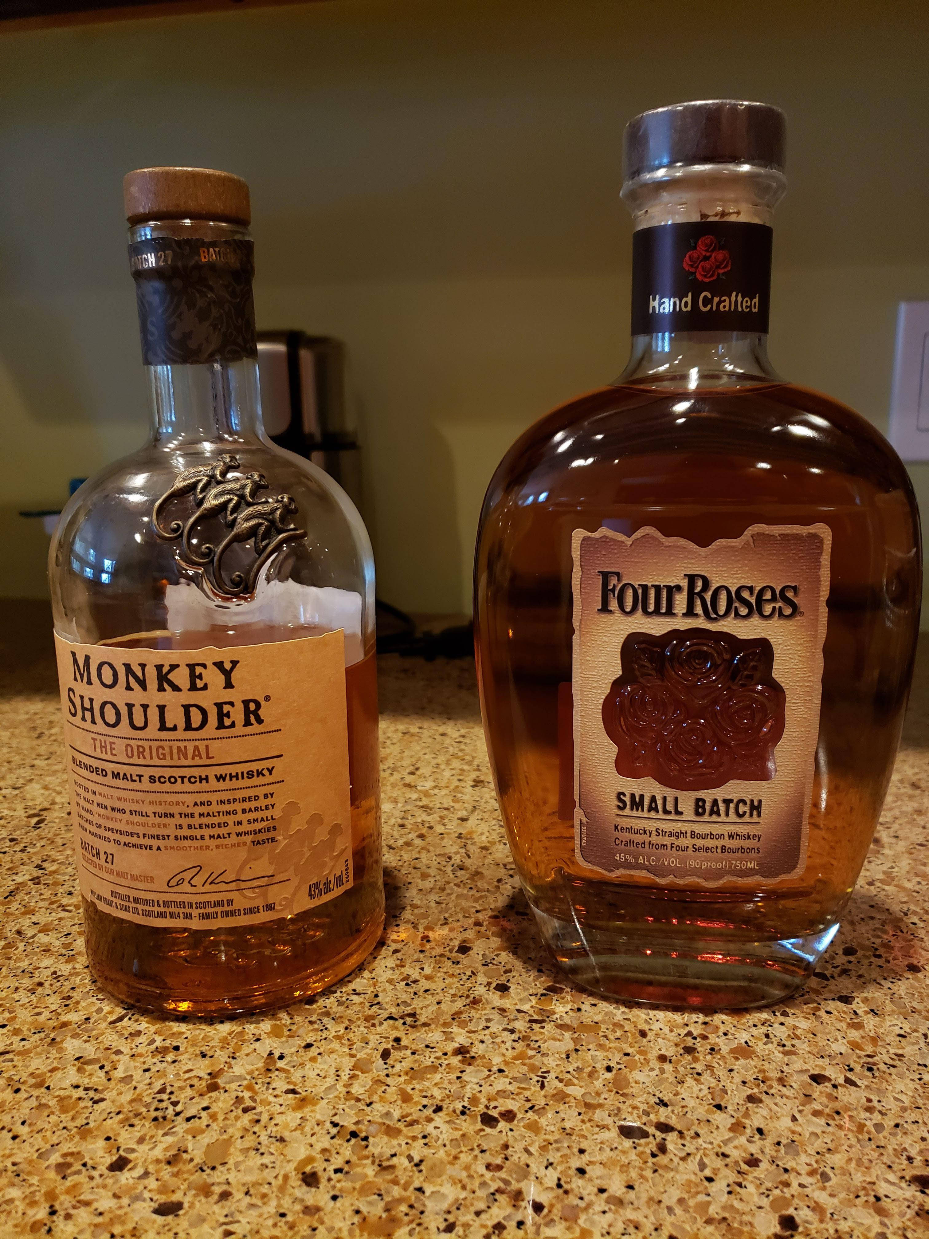 alcohol 桌布 壁紙 — 兩瓶威士忌Monkey Shoulder和Four Roses並排放在有斑點的檯面上，在溫暖的室內燈光下。