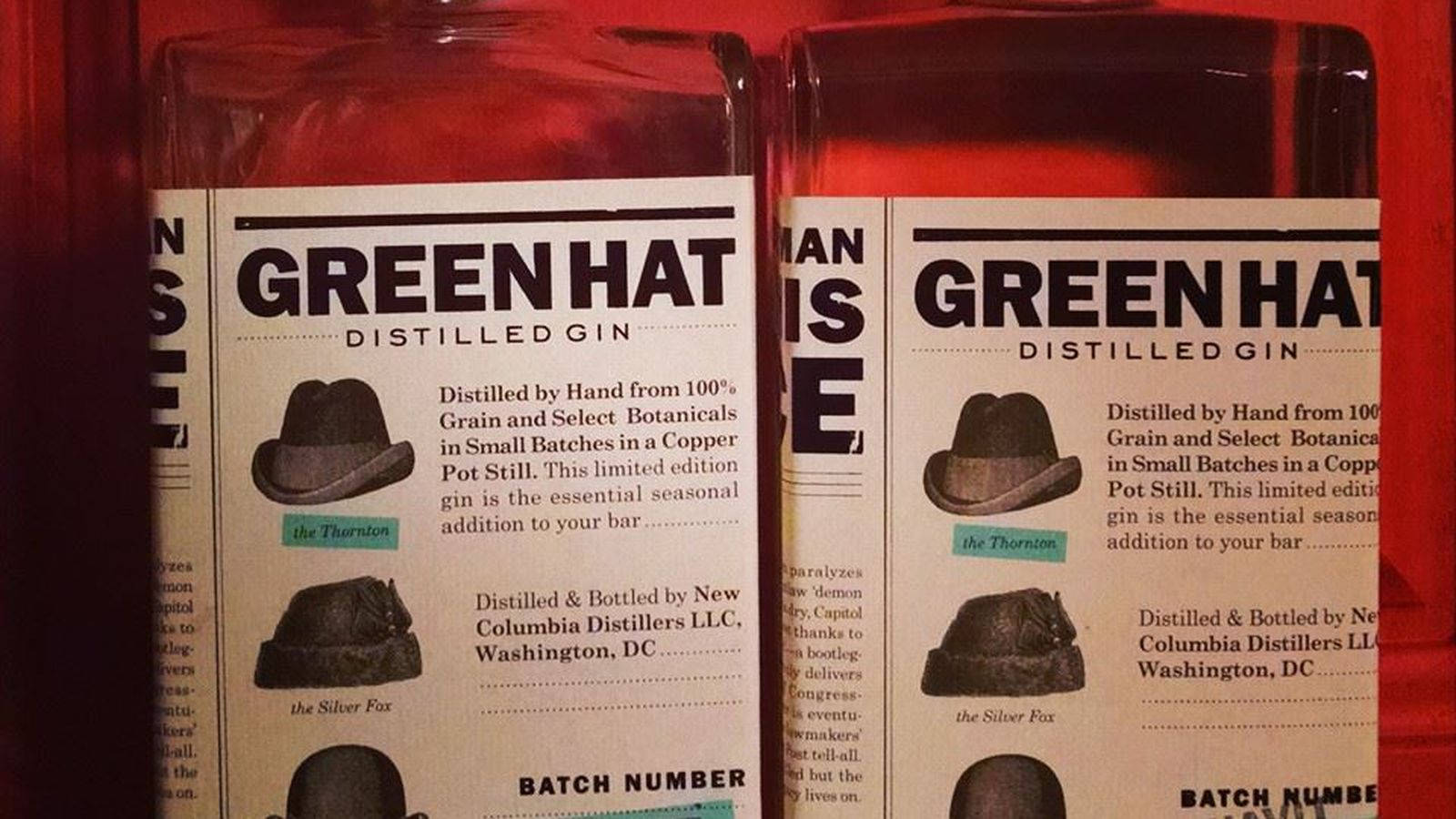 Gin 桌布 壁紙 — 兩瓶帶有帽子插圖復古標籤的 Green Hat 蒸餾琴酒並排放置在深紅色背景前。