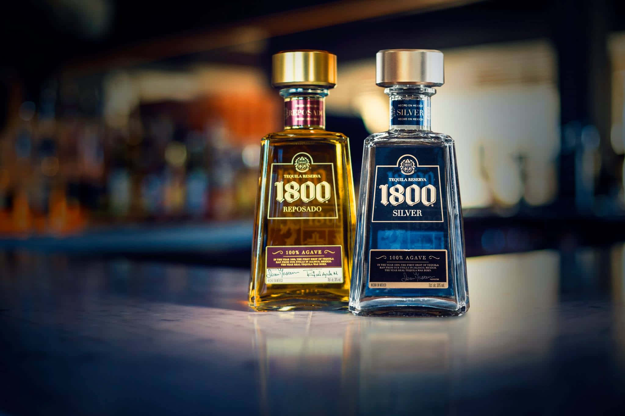 alcohol 桌布 壁紙 — 兩瓶琥珀色和銀色的1800龍舌蘭酒放在拋光吧檯上，背後是模糊的酒架。