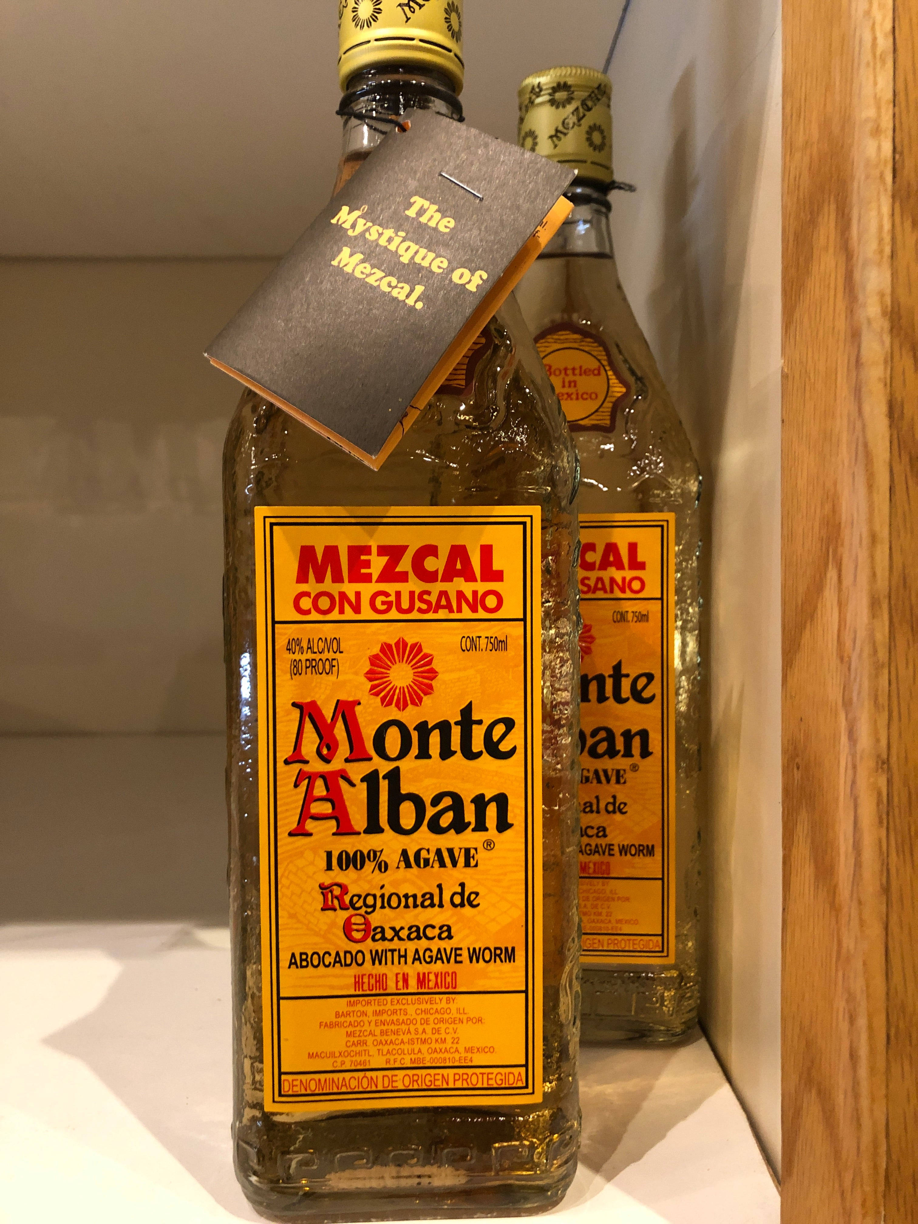 Drink 桌布 壁紙 — 兩瓶琥珀色梅斯卡爾酒，帶有鮮明的黃色標籤，放在架子上，其中一瓶標有「The Mystique of Mezcal」，背景為木質。