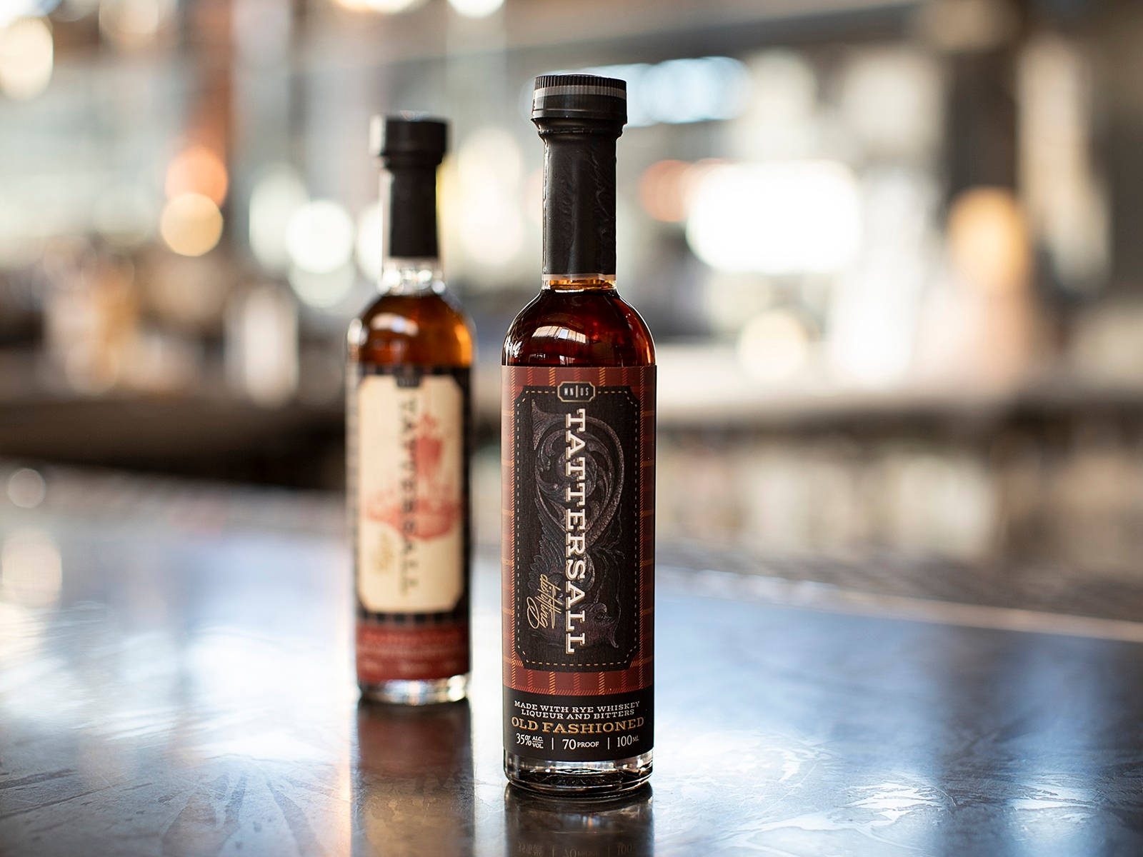 cocktail 桌布 壁紙 — 兩瓶琥珀色的Tattersall Old Fashioned糖漿放在反射的吧檯上，背景有模糊的燈光。