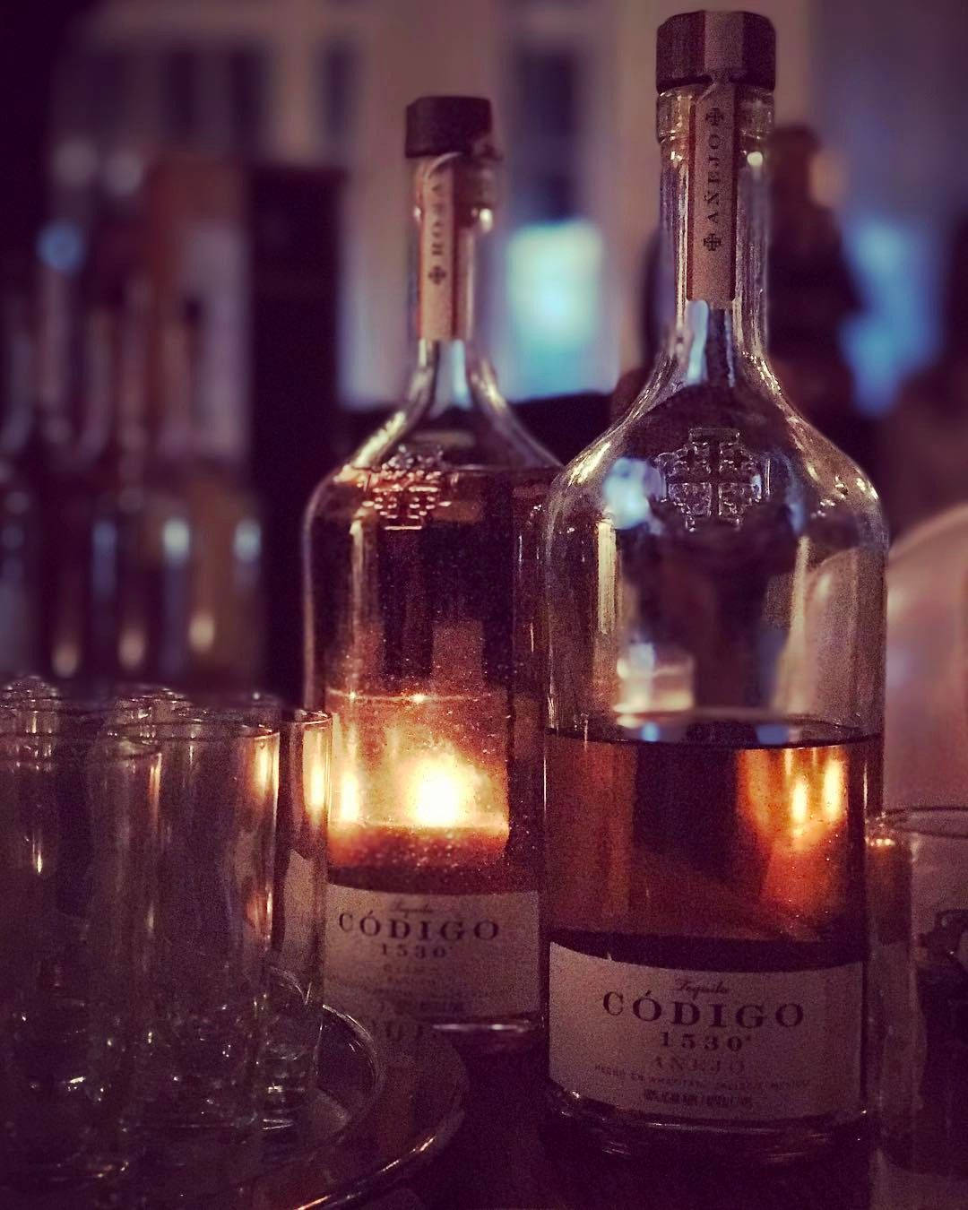 bottle 桌布 壁紙 — 兩瓶發光的 Codigo 1530 龍舌蘭酒放在吧檯上，燭光在玻璃表面反射。