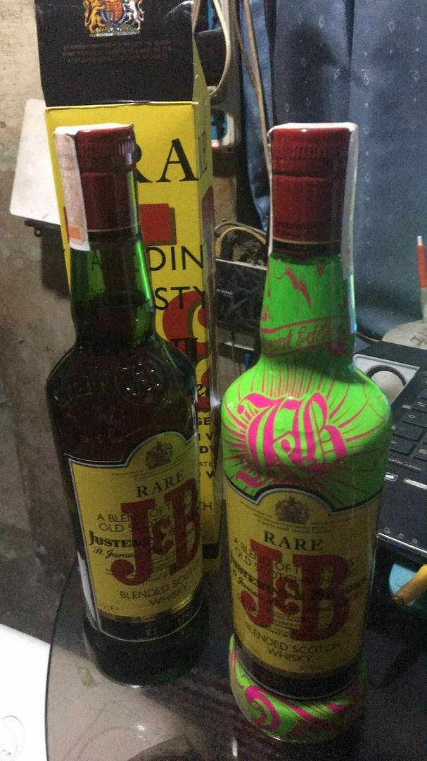 drinks 桌布 壁紙 — 兩瓶罕見的J&B威士忌酒瓶，一瓶標準版，一瓶定制塗成綠色和粉紅色，放在玻璃桌上，旁邊有一台筆記型電腦和雜亂的背景。