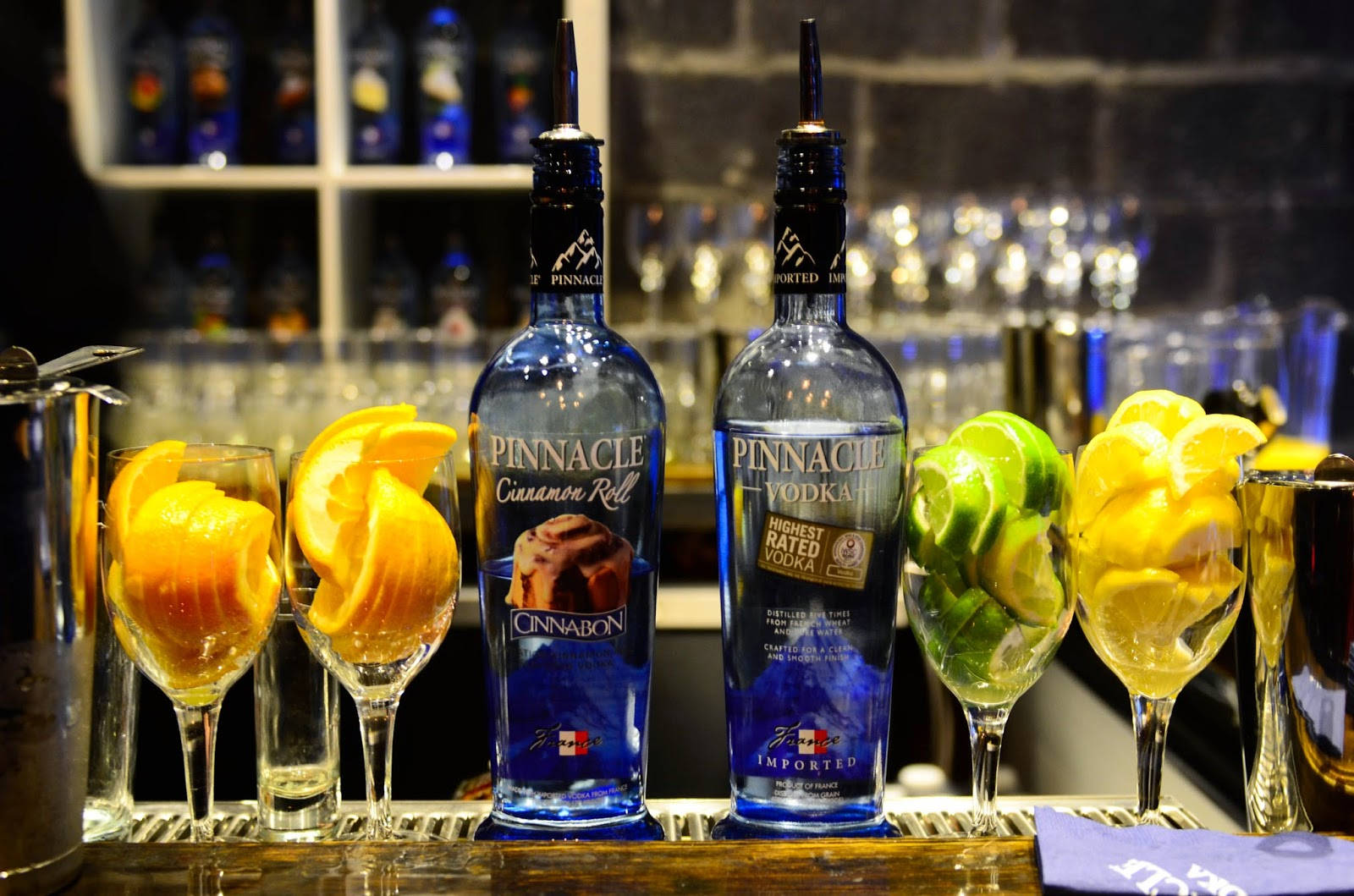 bottle 桌布 壁紙 — 兩瓶藍色Pinnacle伏特加酒瓶夾著裝滿柑橘片的玻璃杯，放在燈光昏暗的酒吧檯上。