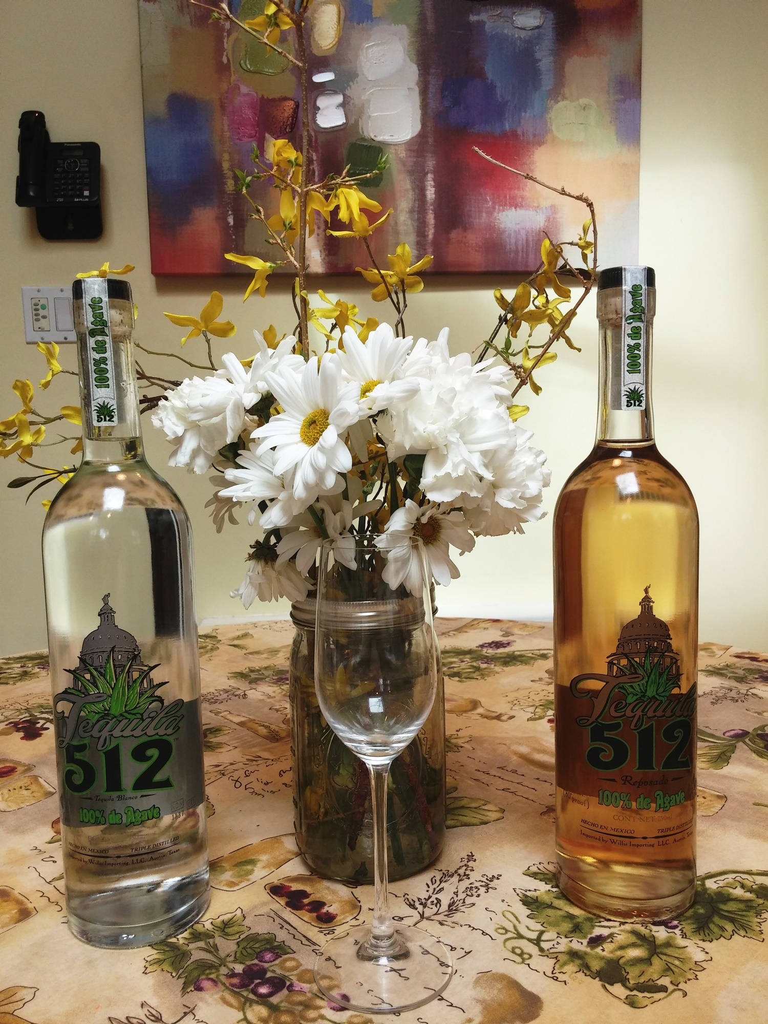 drinks 桌布 壁紙 — 兩瓶龍舌蘭酒放在花桌布上，中間夾著一瓶白色雛菊和黃色連翹，在溫暖的室內燈光下。