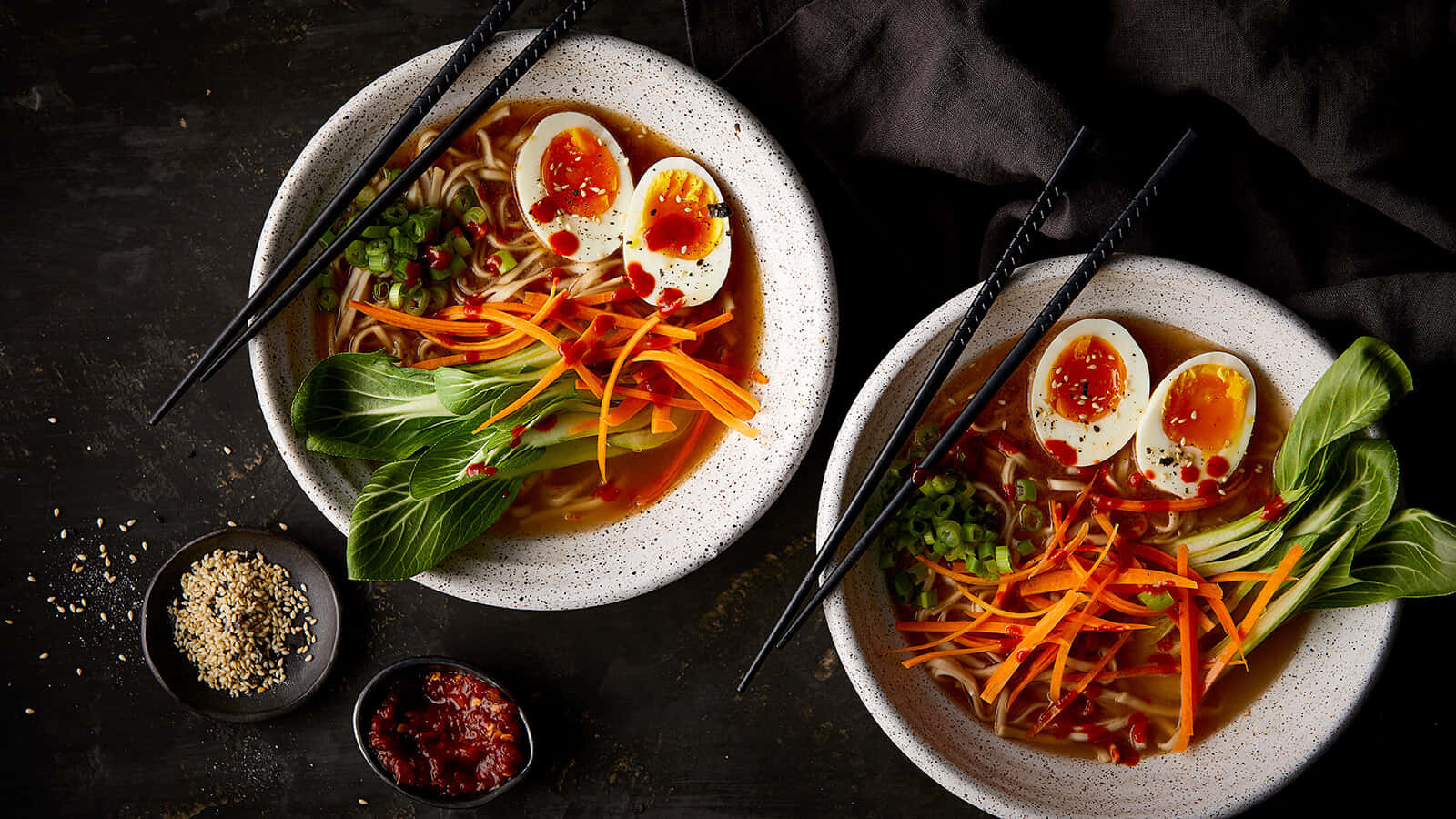 Noodles 桌布 壁紙 — 兩碗冒煙的拉麵，上面放著半熟蛋、切絲紅蘿蔔和青江菜，放在深色質樸的表面上。