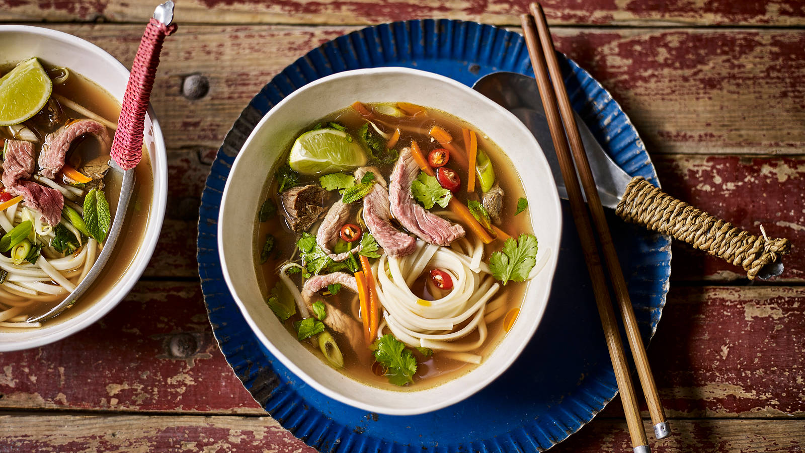 Noodles 桌布 壁紙 — 兩碗冒著熱氣的越南河粉，配有牛肉麵、青檸、香草和辣椒，放在一張質樸的紅色木桌上。