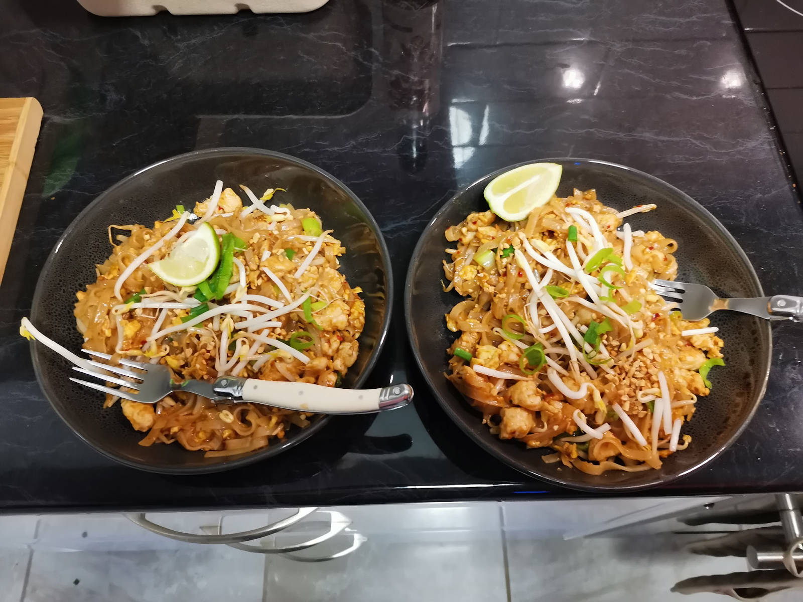 thai 桌布 壁紙 — 兩碗熱騰騰的泰式炒河粉，上面放有萊姆角、豆芽和青蔥，放在光滑的黑色檯面上。
