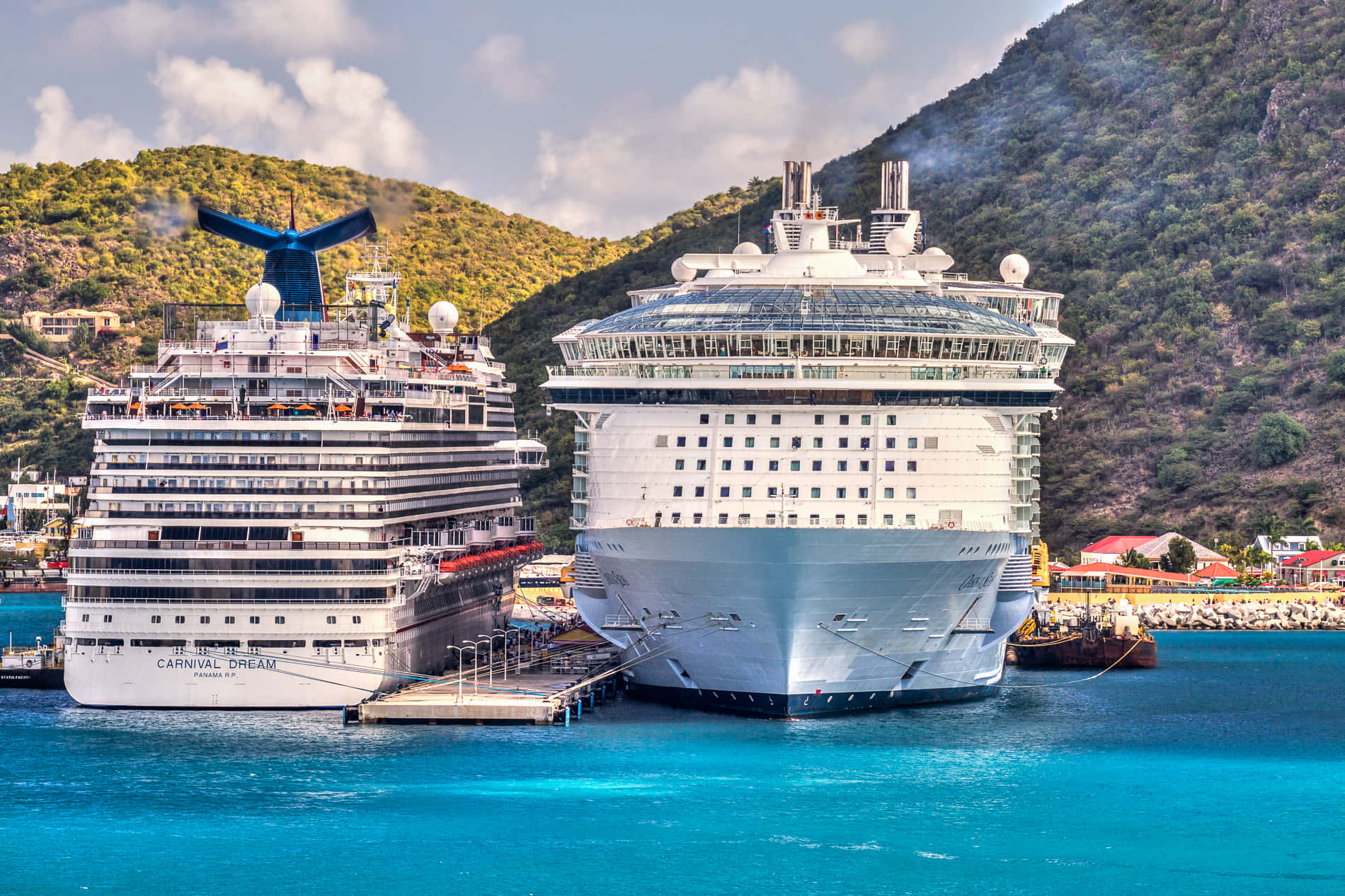 Carnival Dream 桌布 壁紙 — 兩艘巨型郵輪並排停泊在碧綠色的海水中，背後有綠色山丘在部分多雲的天空下聳立。