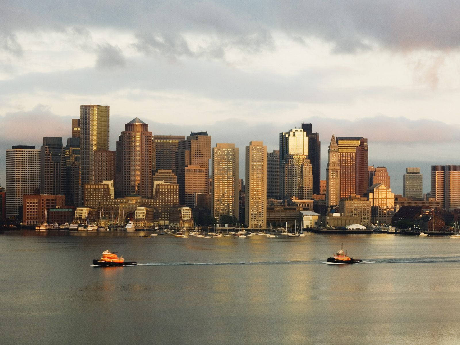 Boston 桌布 壁紙 — 兩艘橙色拖船在平靜的水面上滑行，前方是沐浴在金色日落光芒中的高聳城市天際線。