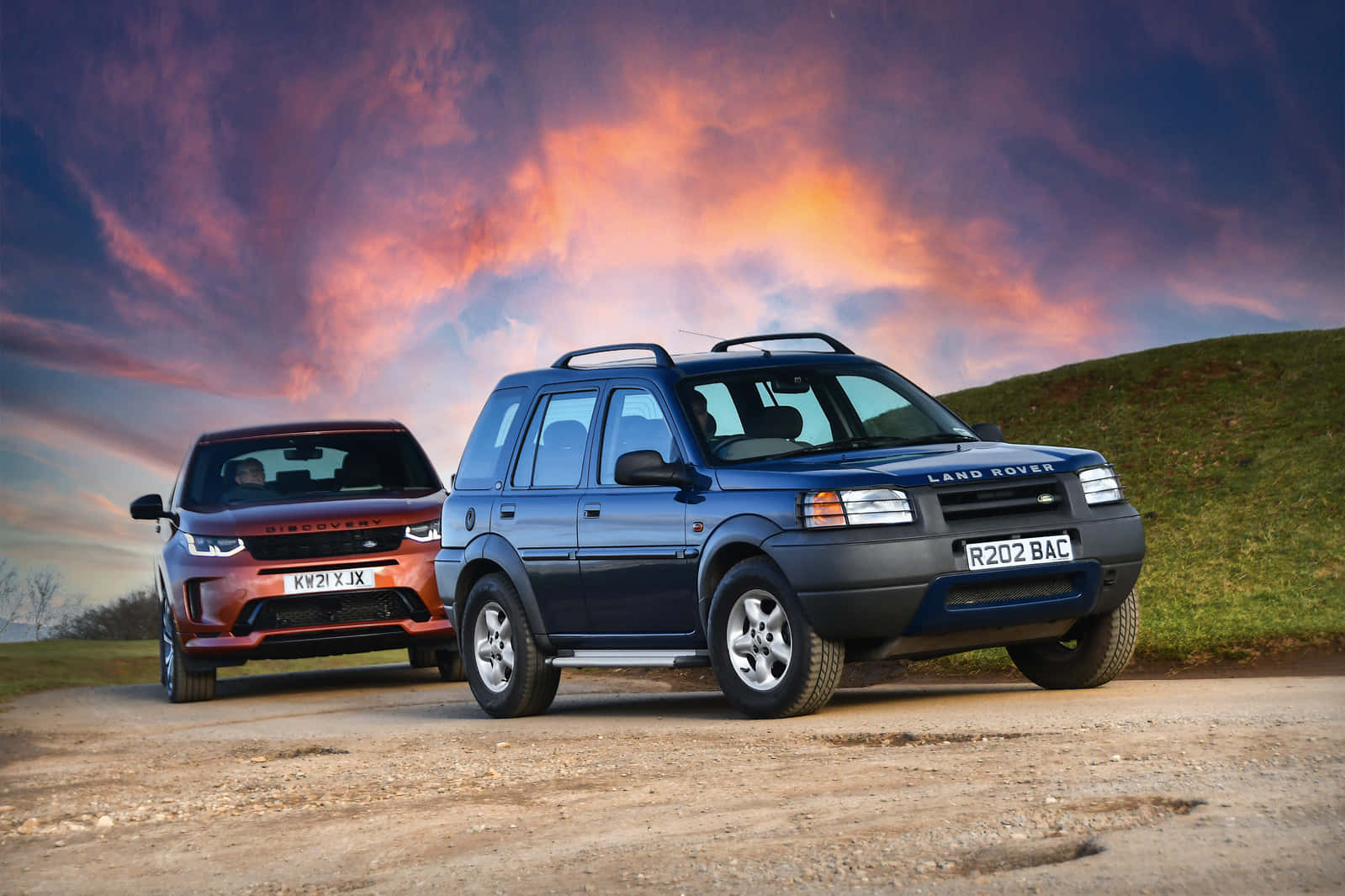 Land Rover Freelander 桌布 壁紙 — 兩輛 Land Rover Discovery SUV 停在一條土路上，背景是壯麗的日落天空，帶有火橙色和紫色的雲彩。