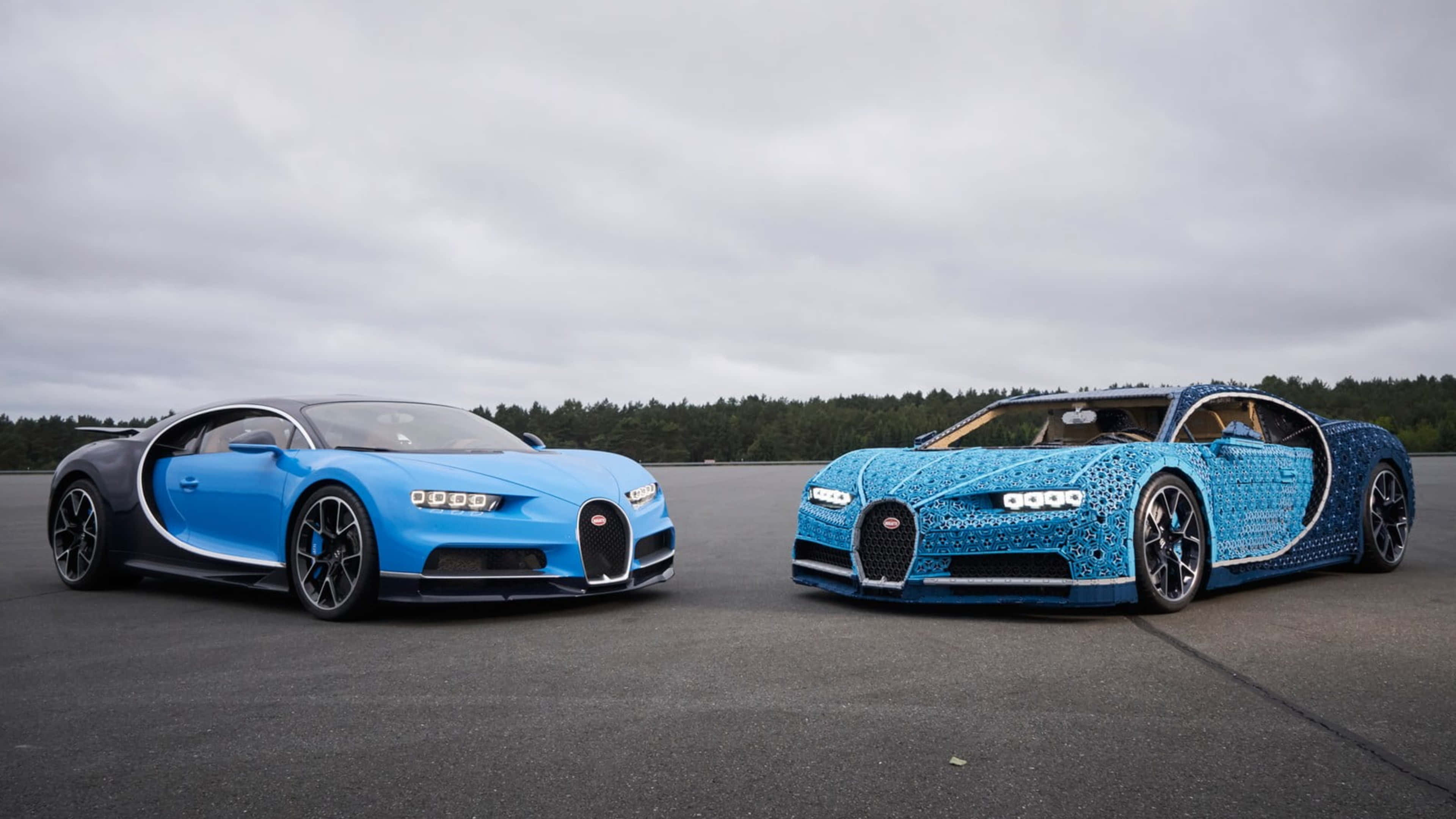 bugatti iPhone 桌布 壁紙 — 兩輛Bugatti Chiron超級跑車，一輛真車和一輛用LEGO積木搭建的，並排停在陰天下的瀝青賽道上。