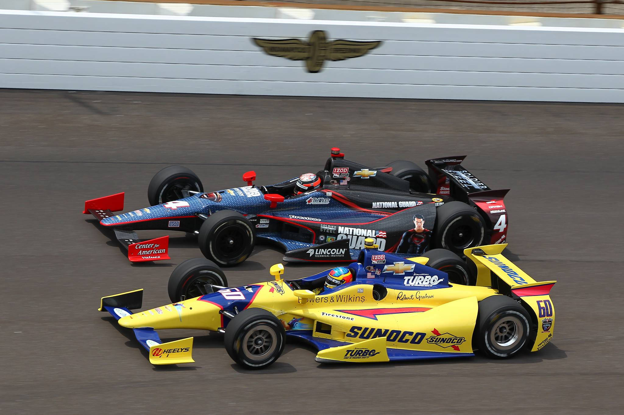 Indianapolis 桌布 壁紙 — 兩輛IndyCar賽車在賽道上並排飛馳，背景牆上有贊助商標誌和帶翼標誌。