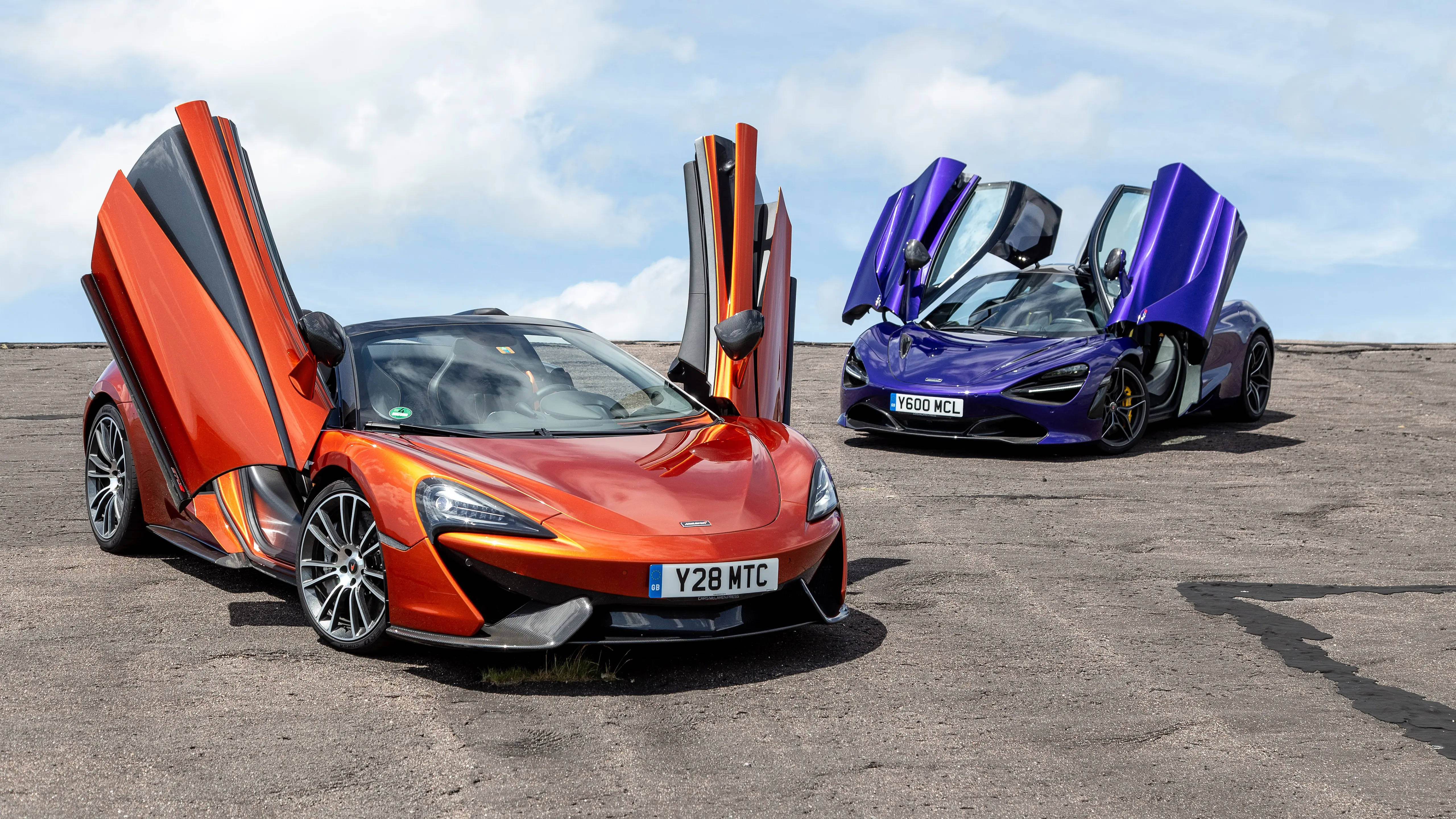 跑車 桌布 壁紙 — 兩輛McLaren跑車打開鷗翼車門，一橙一紫，停泊在碎石地上，天空部分多雲。