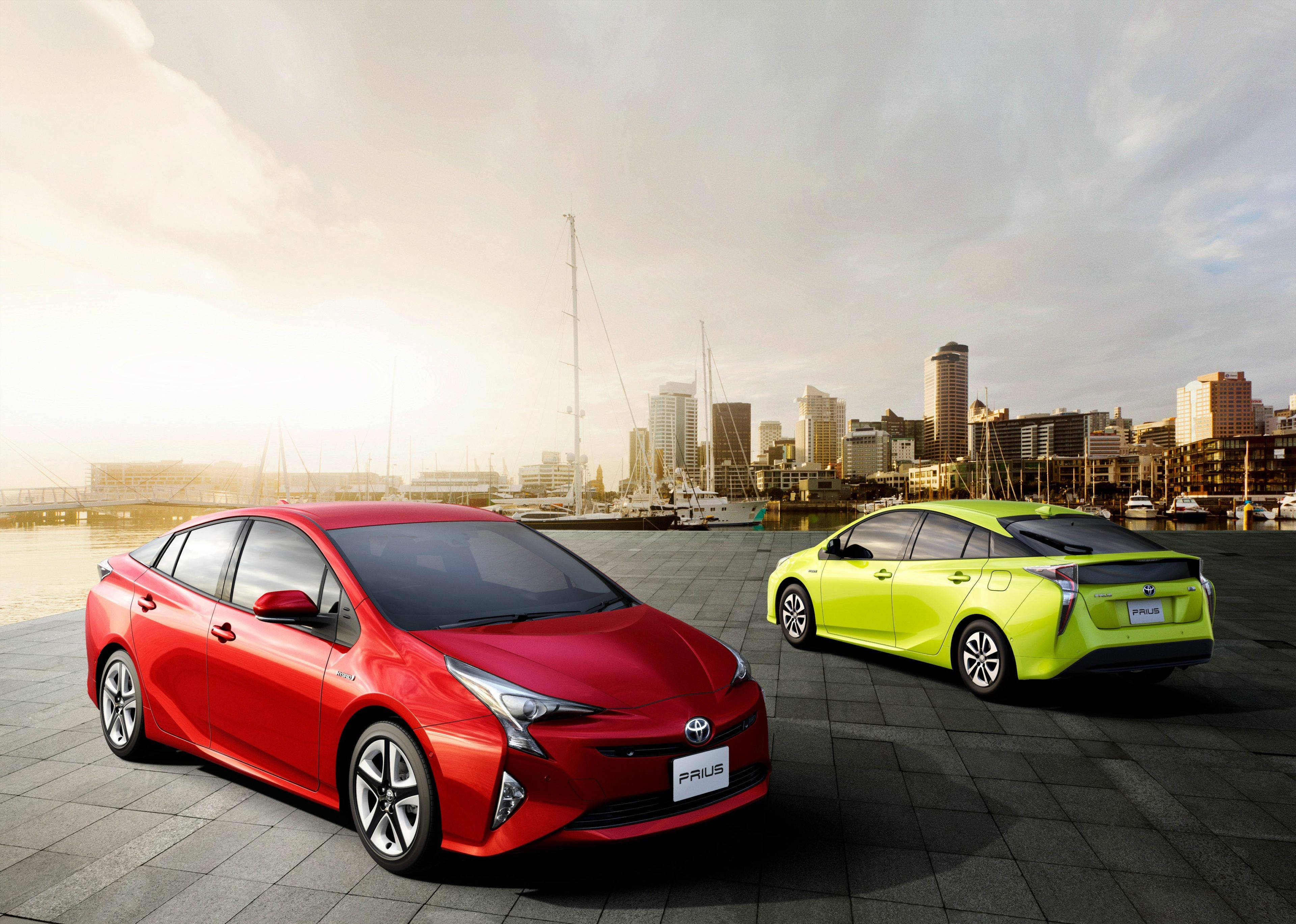 Toyota 桌布 壁紙 — 兩輛充滿活力的豐田Prius汽車，一輛紅色，一輛萊姆綠色，停在海濱碼頭上，背景是城市天際線和船隻，正值黃金時刻。