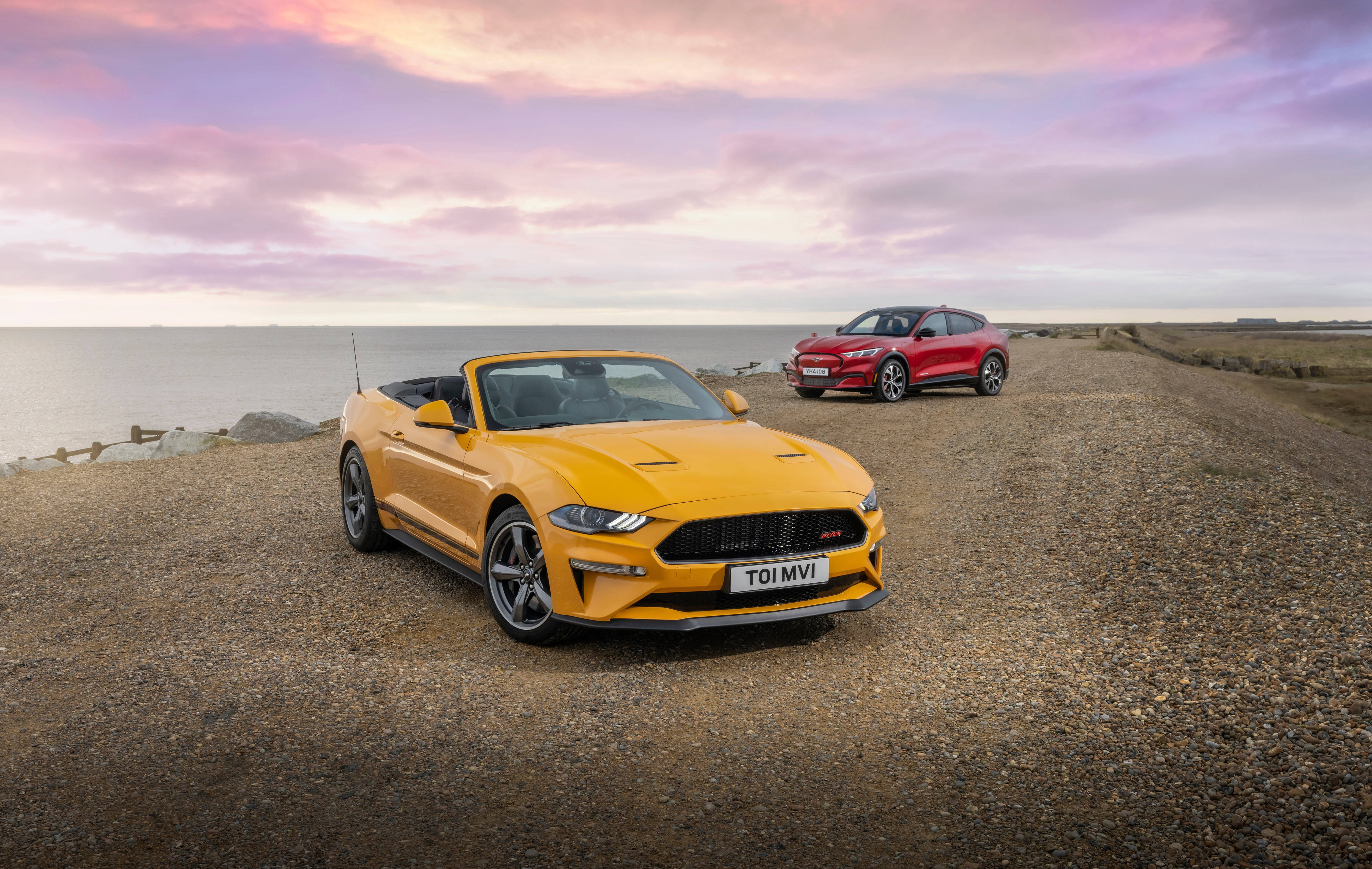Cool Mustang 桌布 壁紙 — 兩輛充滿活力的跑車，一輛黃色敞篷車和一輛紅色轎跑車，停在碎石海岸公路上，背景是粉彩色的日落天空。
