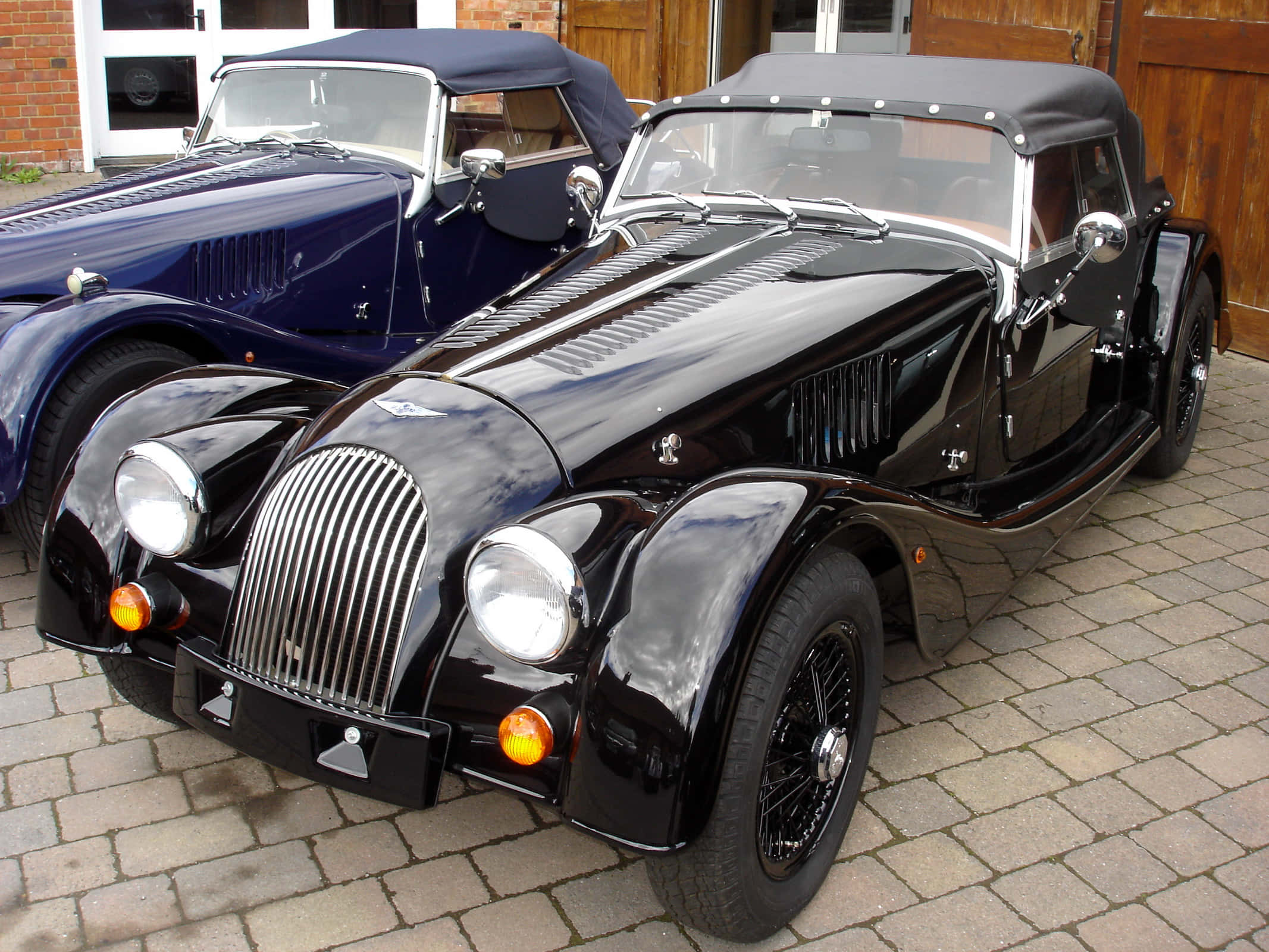Morgan 桌布 壁紙 — 兩輛光澤的復古跑車，帶黑色敞篷車頂，停在磚砌建築附近的石頭庭院上。