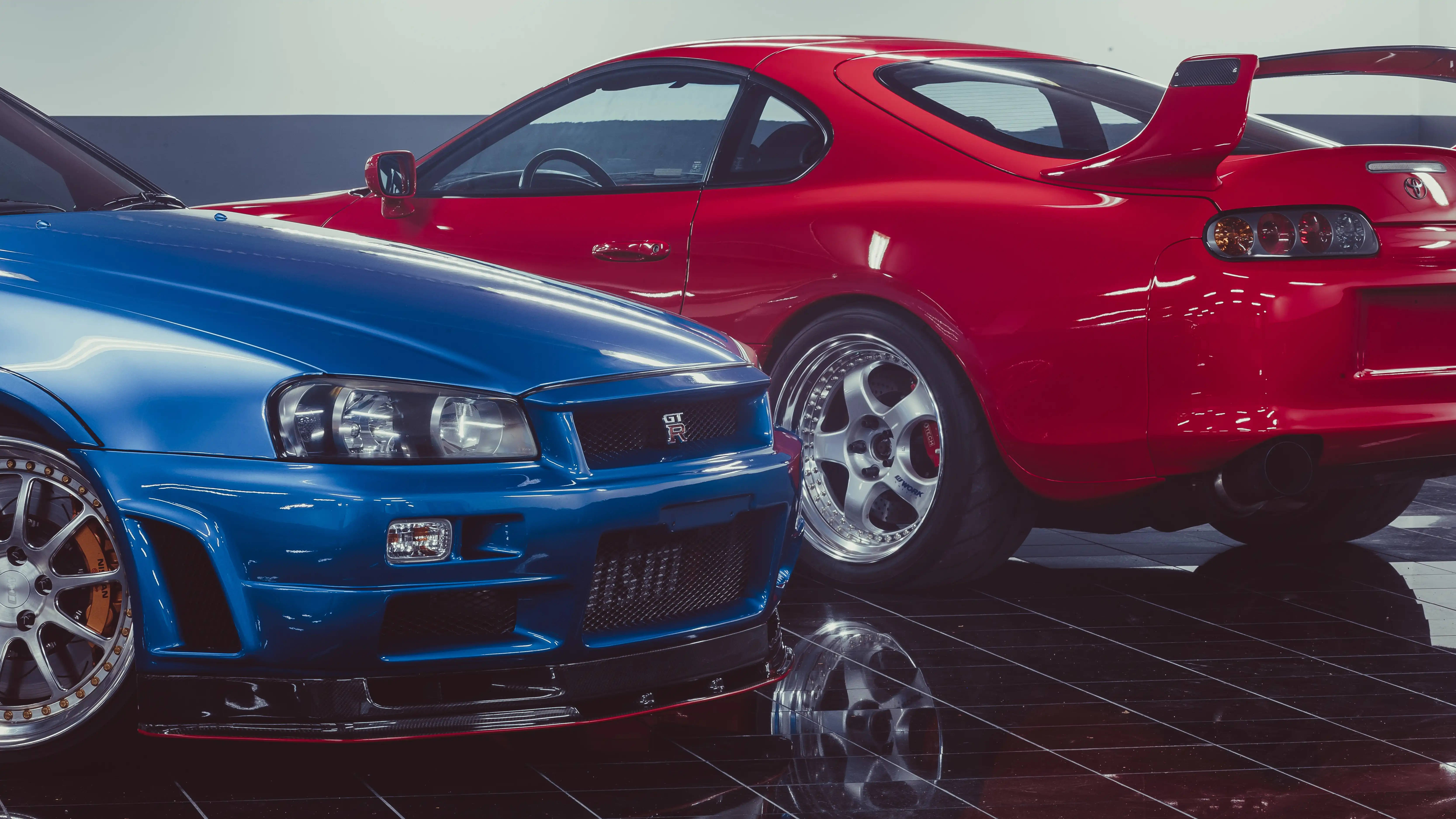 Nissan 桌布 壁紙 — 兩輛光澤跑車，一輛藍色日產GT-R和紅色豐田Supra，停在室內反光黑色瓷磚地板上。