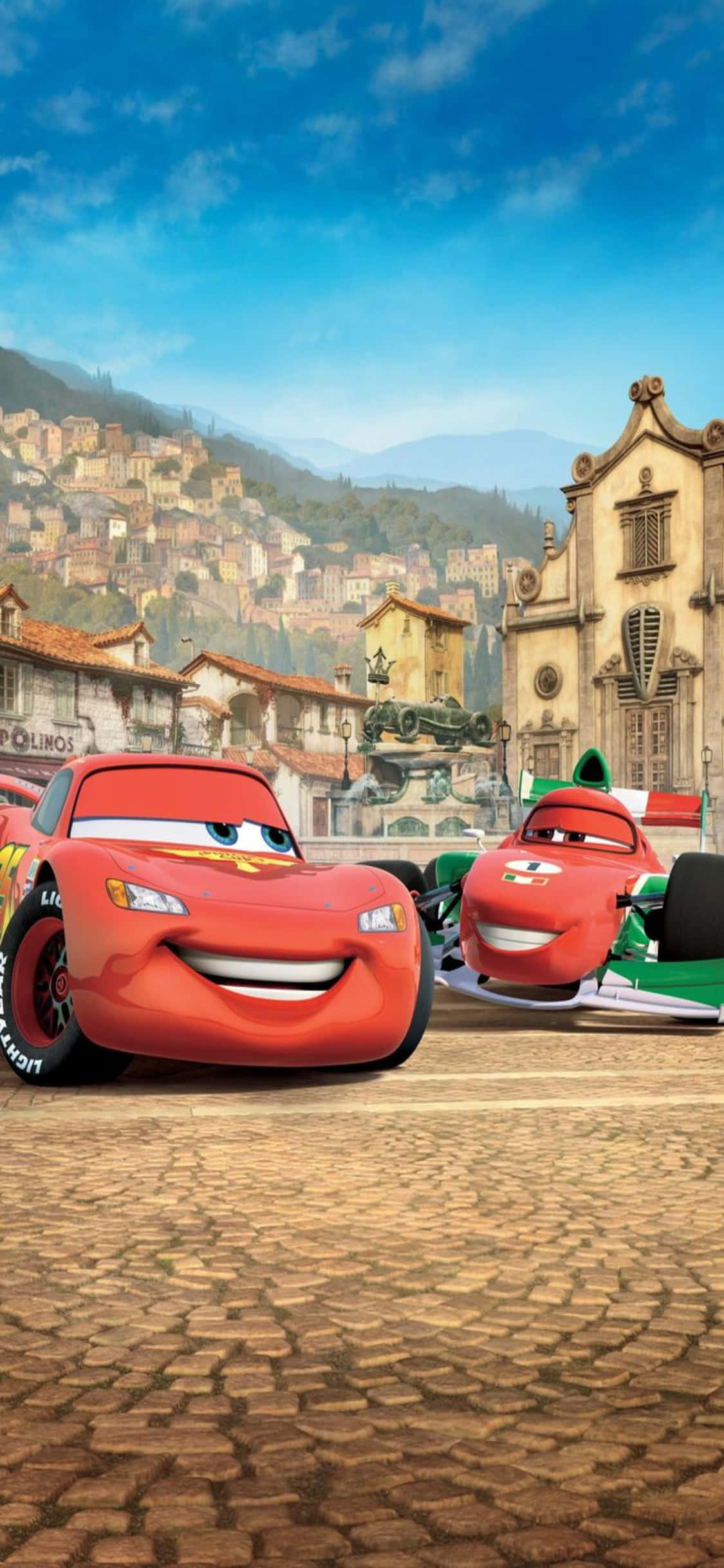 Lightning Mcqueen 桌布 壁紙 — 兩輛動畫賽車在陽光明媚的歐洲小鎮的鵝卵石街道上愉快地微笑，背景是山坡建築。