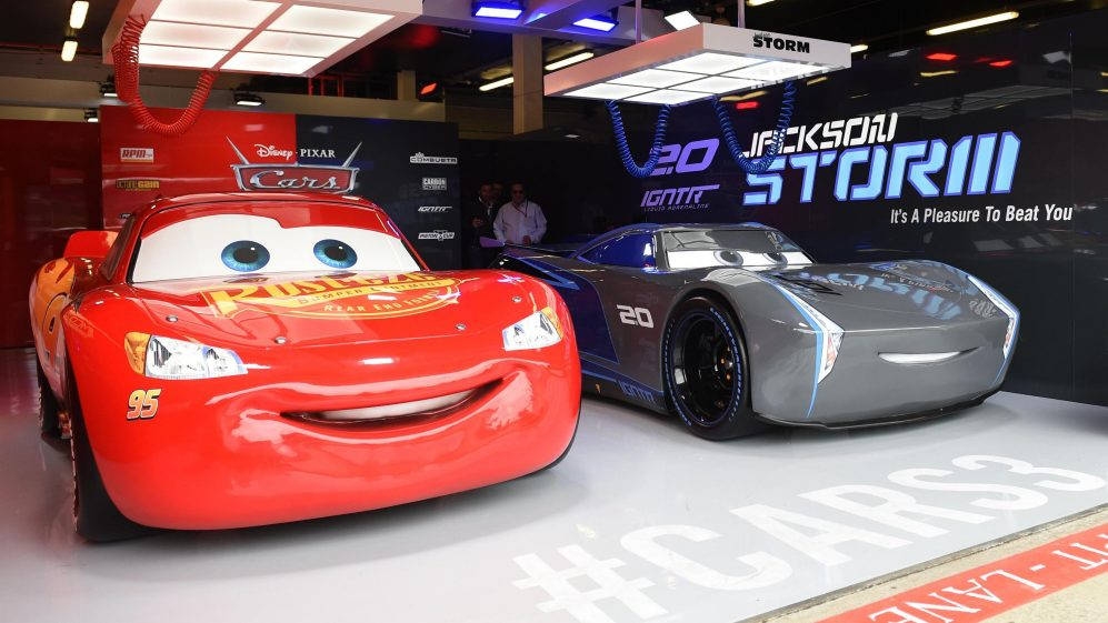 皮克斯 桌布 壁紙 — 兩輛動畫賽車閃電麥坤和傑克森史東並肩站在Cars 3宣傳活動中，周圍有發光標誌和明亮燈光。