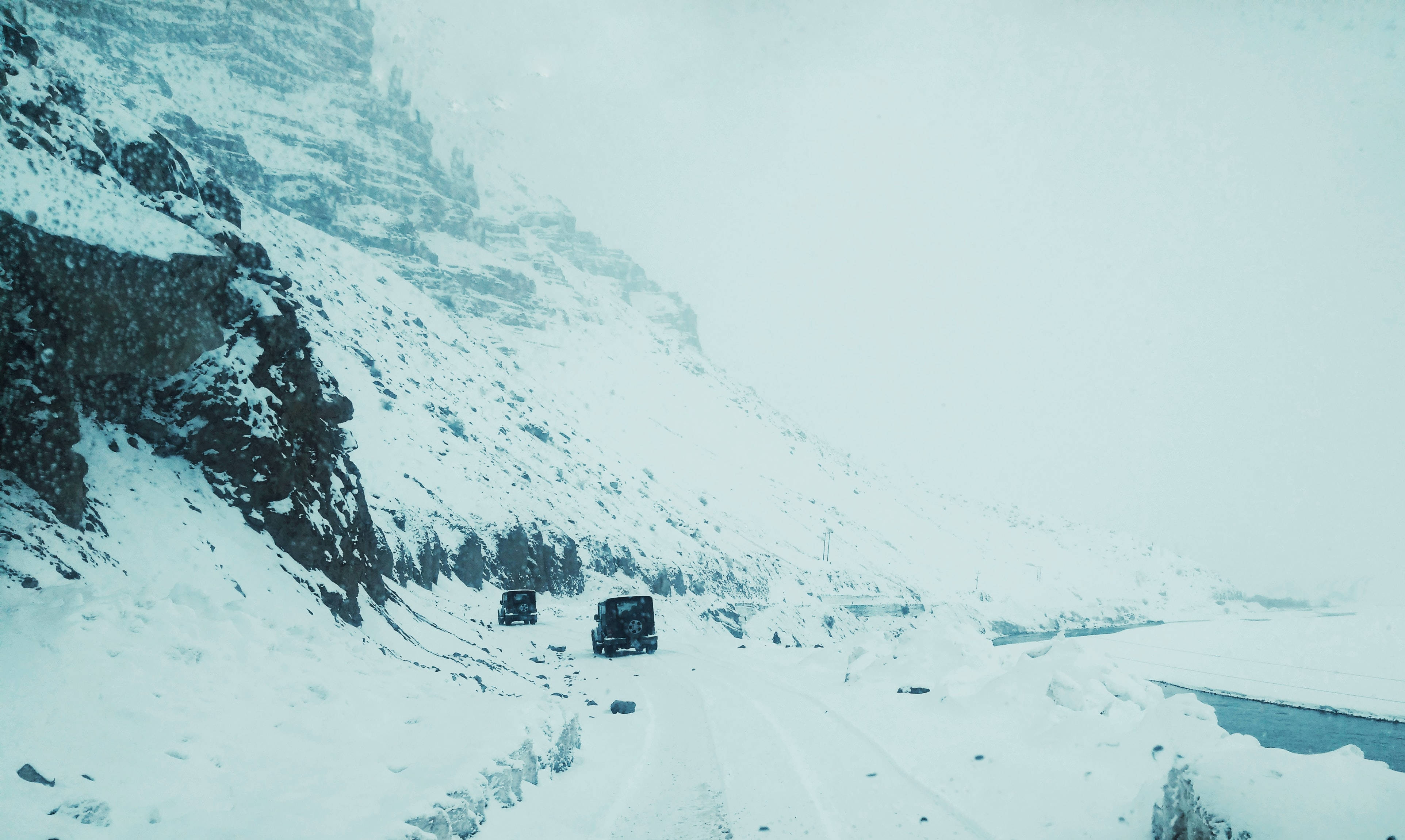 山景 桌布 壁紙 — 兩輛卡車在暴風雪中行駛於積雪的山路上，一側是陡峭的岩石懸崖，下方是一條結冰的河流。