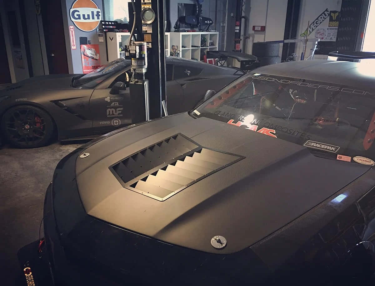 Car Hood 桌布 壁紙 — 兩輛啞光黑色賽車配備碳纖維引擎蓋，停放在海灣加油站標誌下光線昏暗的車庫中。
