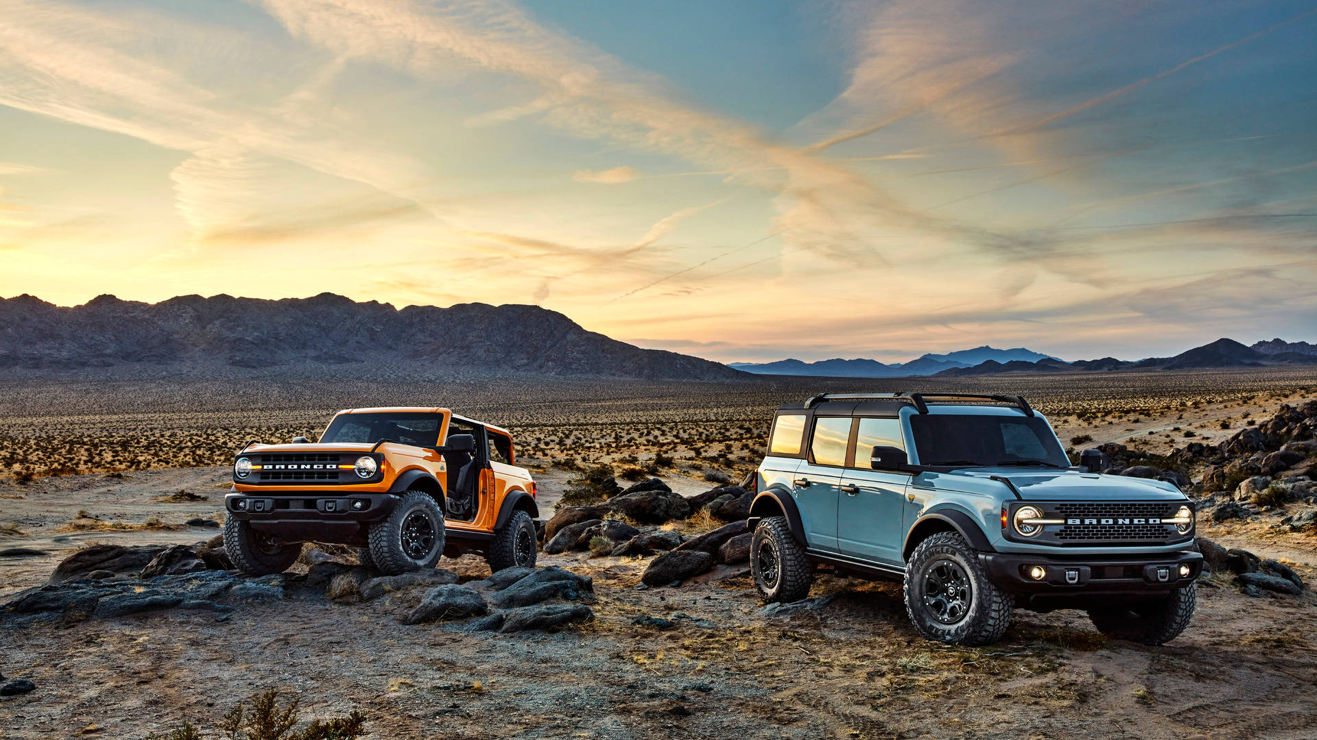 Ford Bronco 桌布 壁紙 — 兩輛堅固的Bronco，一輛橙色一輛藍色，停在岩石沙漠地形上，金色夕陽天空下，遠處有山脈。