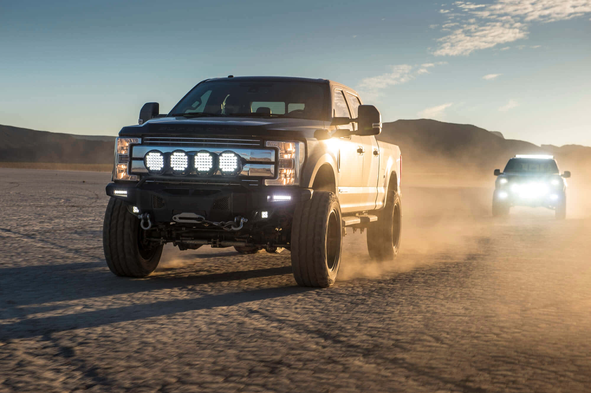 Jacked Up Trucks 桌布 壁紙 — 兩輛堅固的升高卡車在陽光明媚的沙漠乾涸湖床上咆哮而過，在清澈的藍天下揚起灰塵。